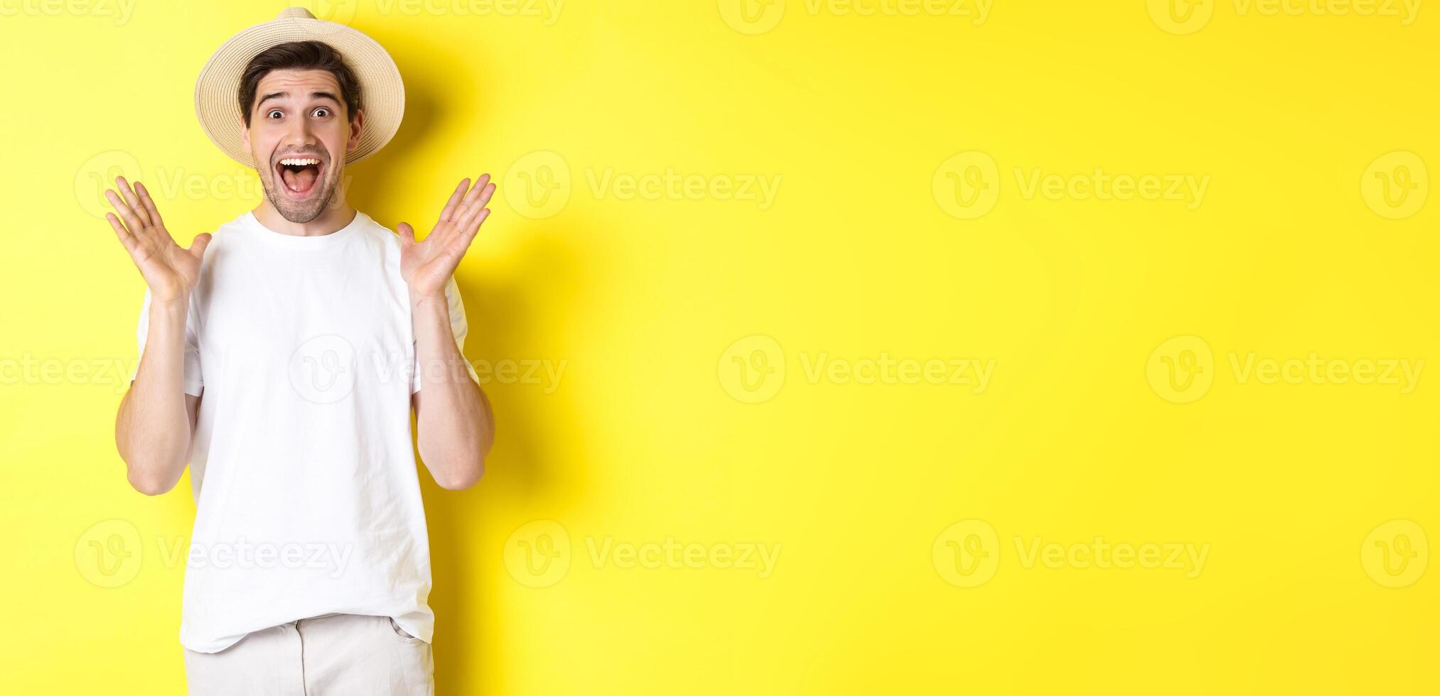Concept of tourism and summer. Happy young man in straw hat looking amazed, reacting to surprise, standing over yellow background photo