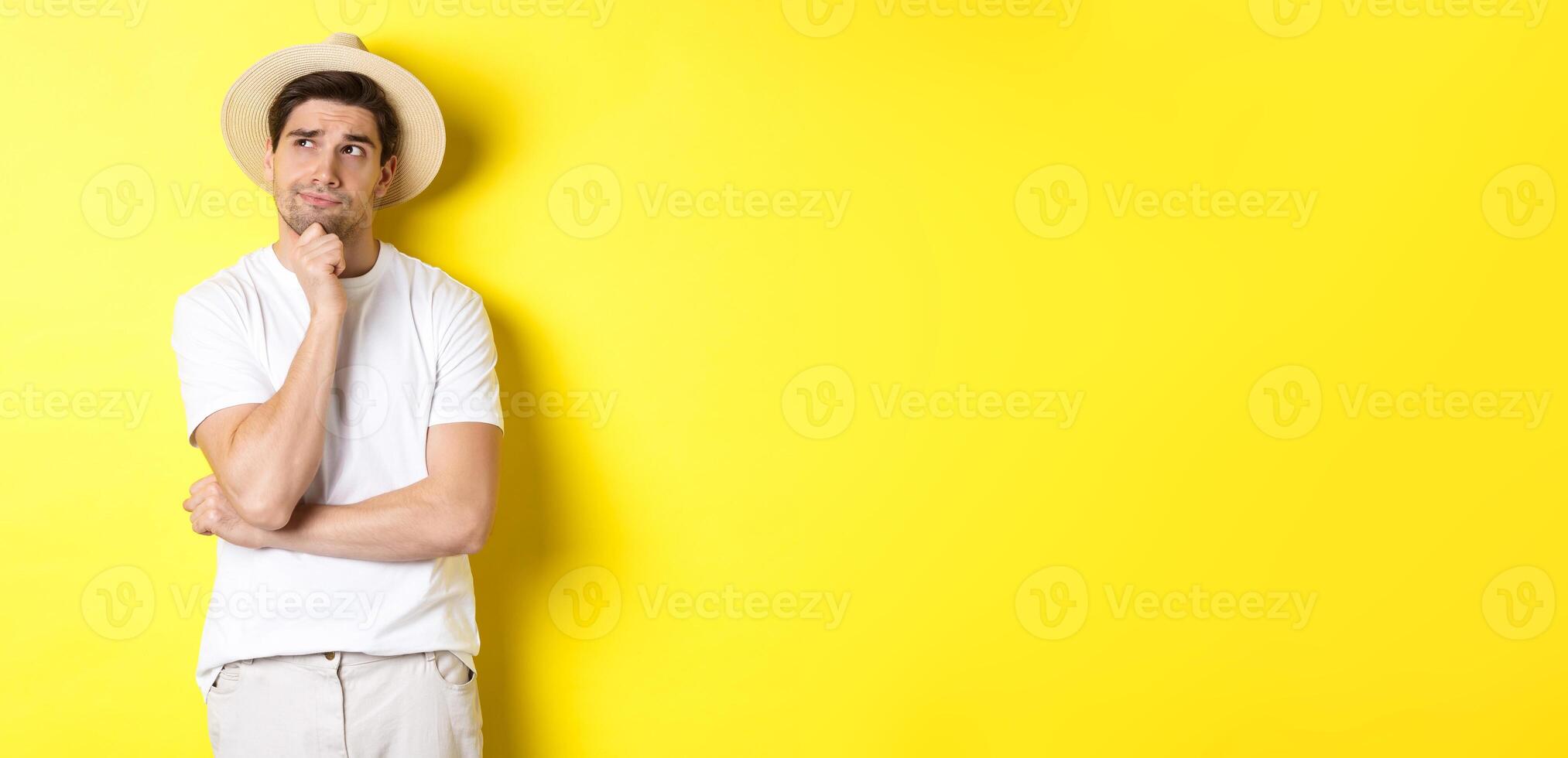 Concept of tourism and summer. Thoughtful man tourist pondering, looking at upper left corner and thinking, standing in straw hat and white t-shirt against yellow background photo