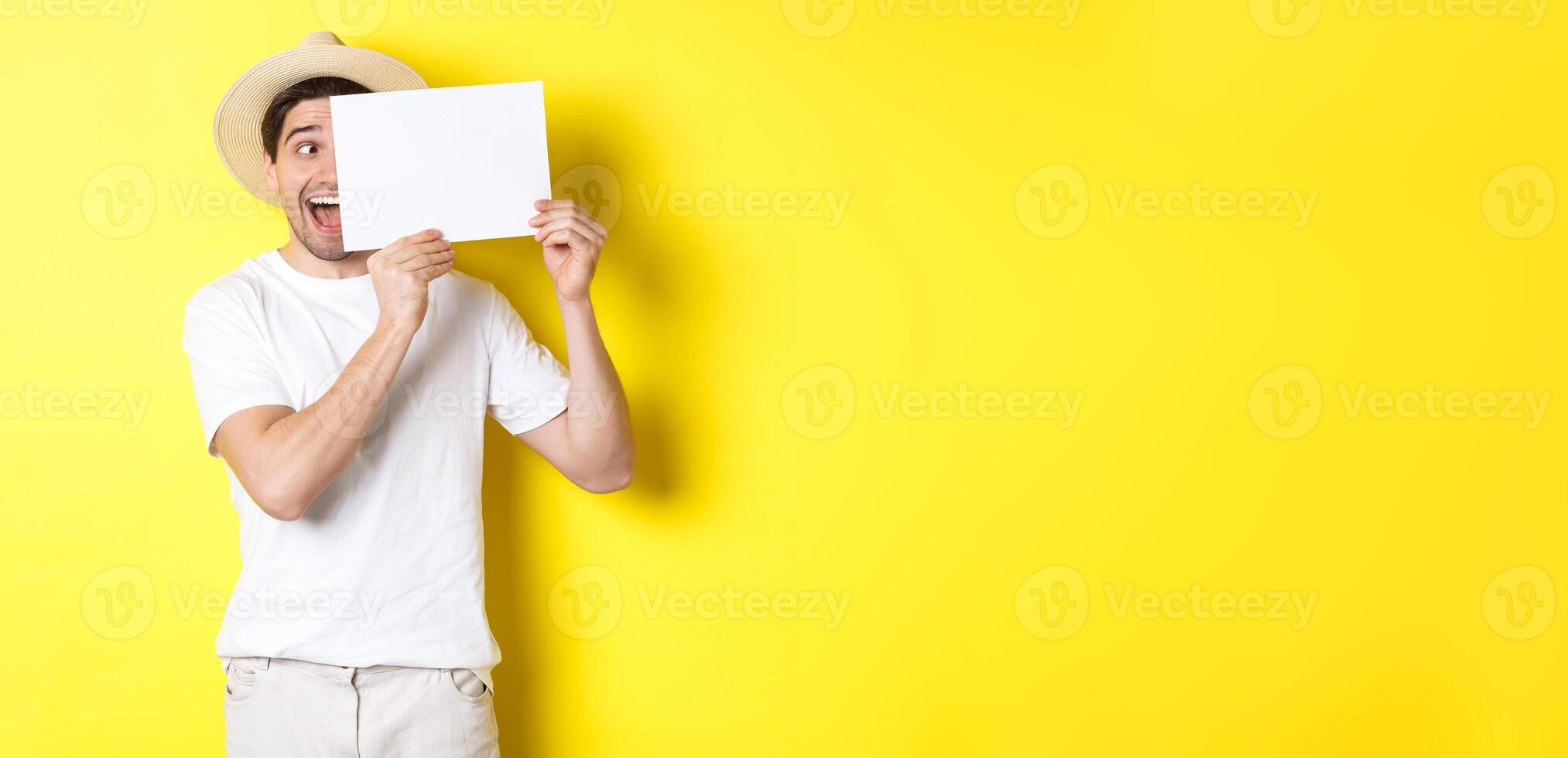 Excited man on vacation showing blank piece of paper for your logo, holding sign near face and smiling, standing against yellow background photo