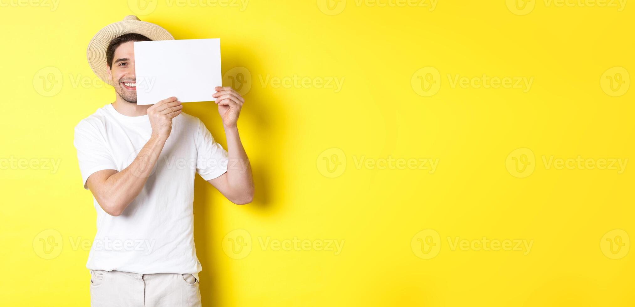 Happy guy on vacation showing blank piece of paper for your logo, holding sign near face and smiling, standing against yellow background photo