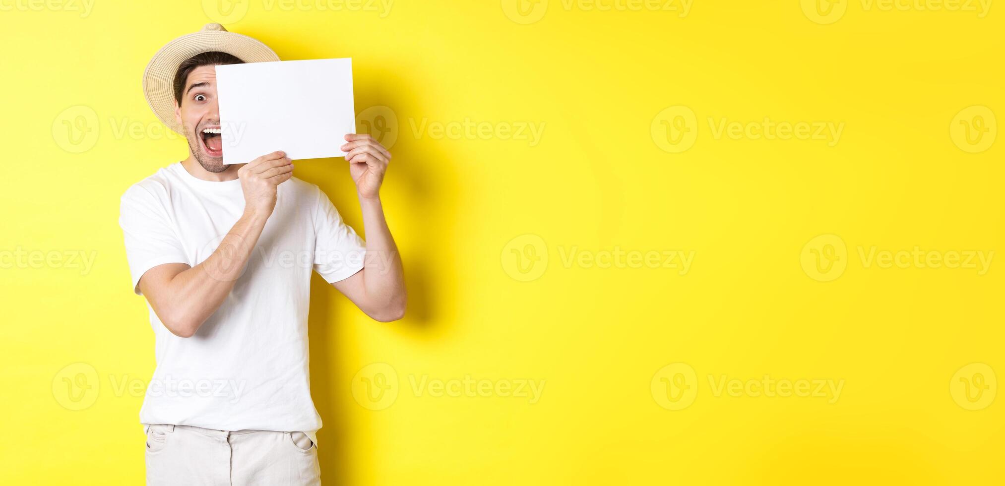 Excited tourist on vacation showing blank piece of paper for your logo, holding sign near face and smiling, standing against yellow background photo