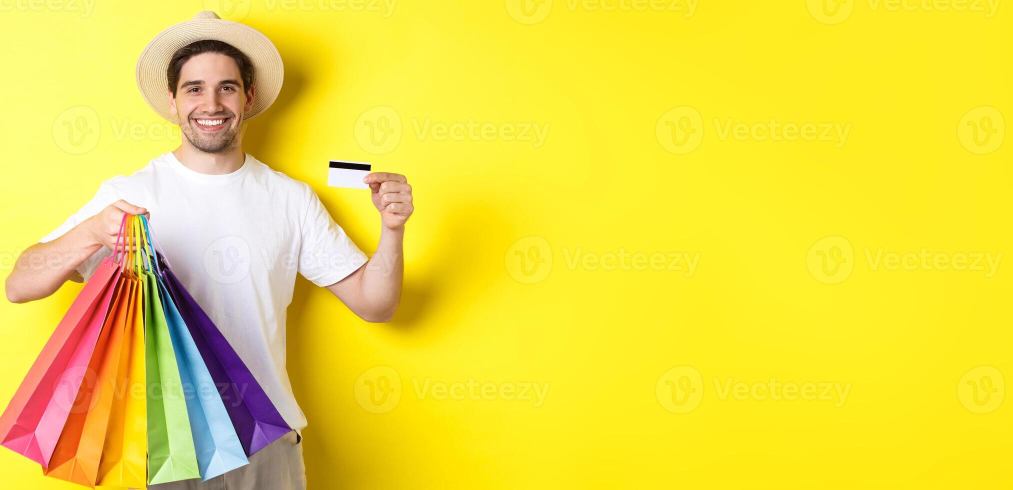 Happy attractive guy showing shopping bags and credit card, concept of banking and easy payment, standing over yellow background photo