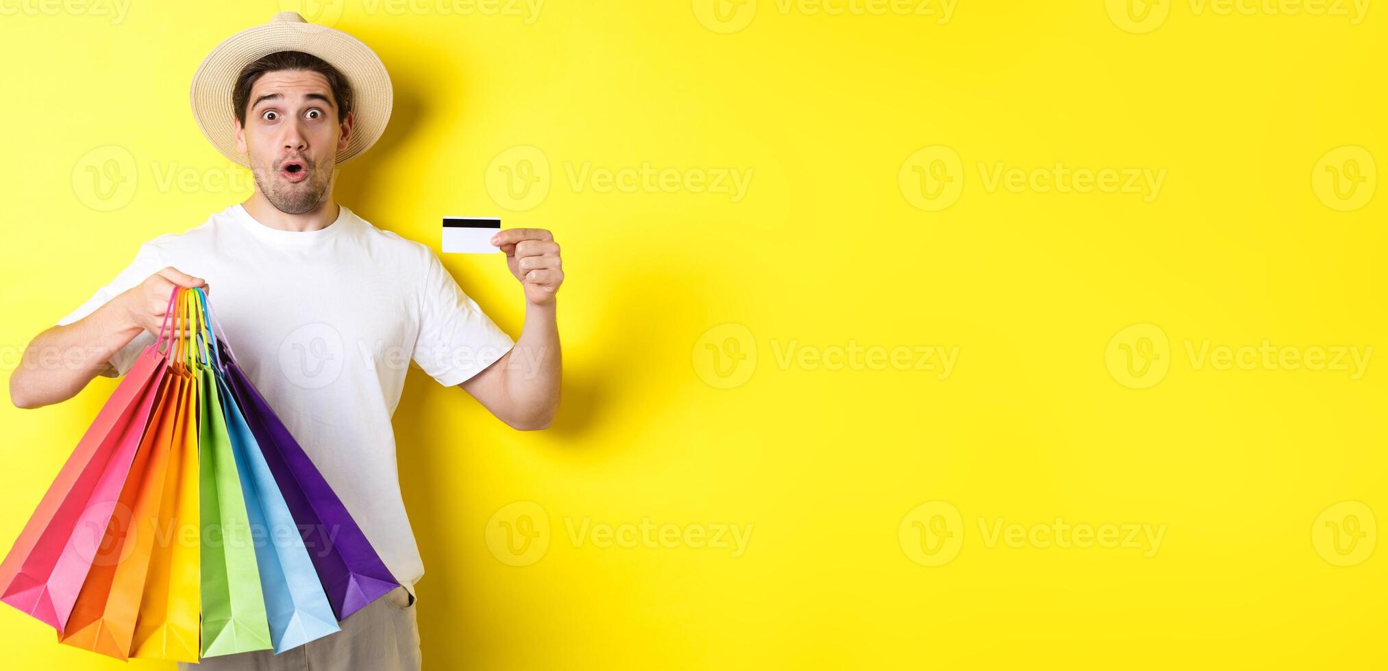 Impressed man showing shopping bags with products and credit card, standing over yellow background photo