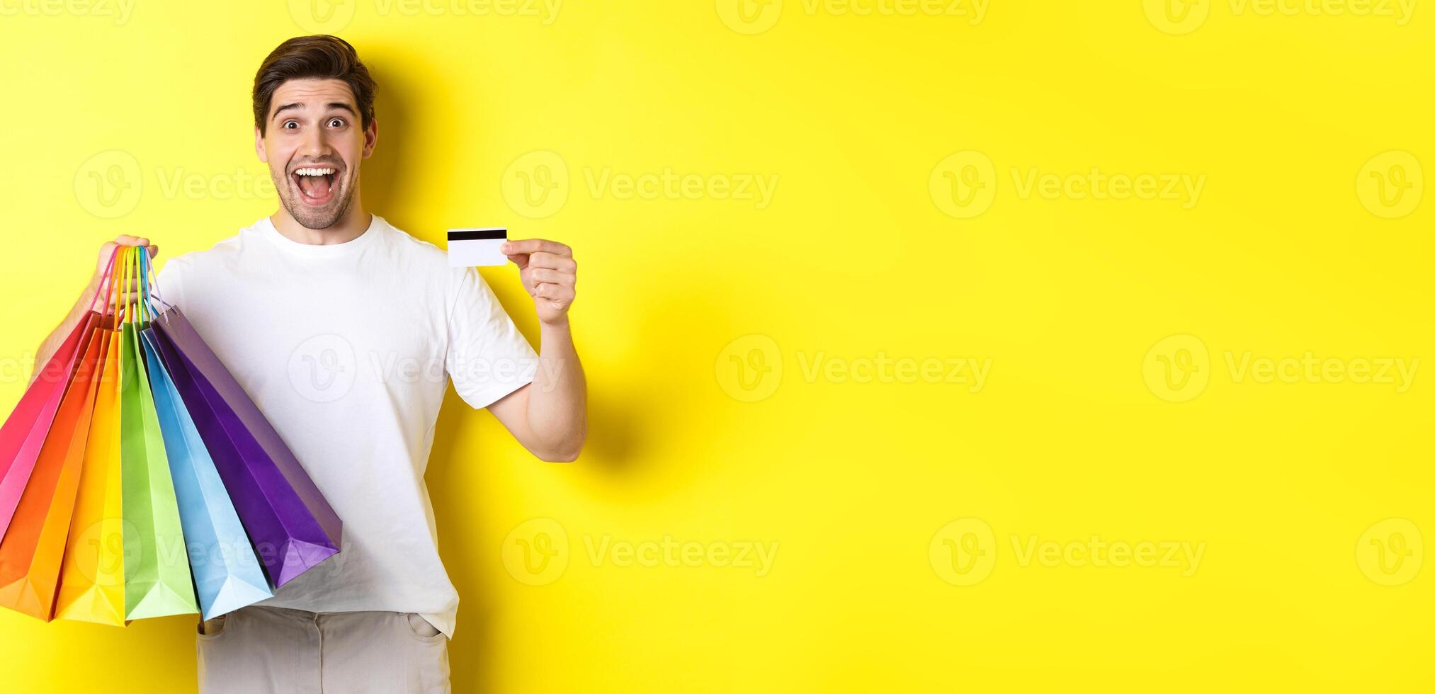 Happy attractive man holding shopping bags, showing credit card, standing over yellow background photo