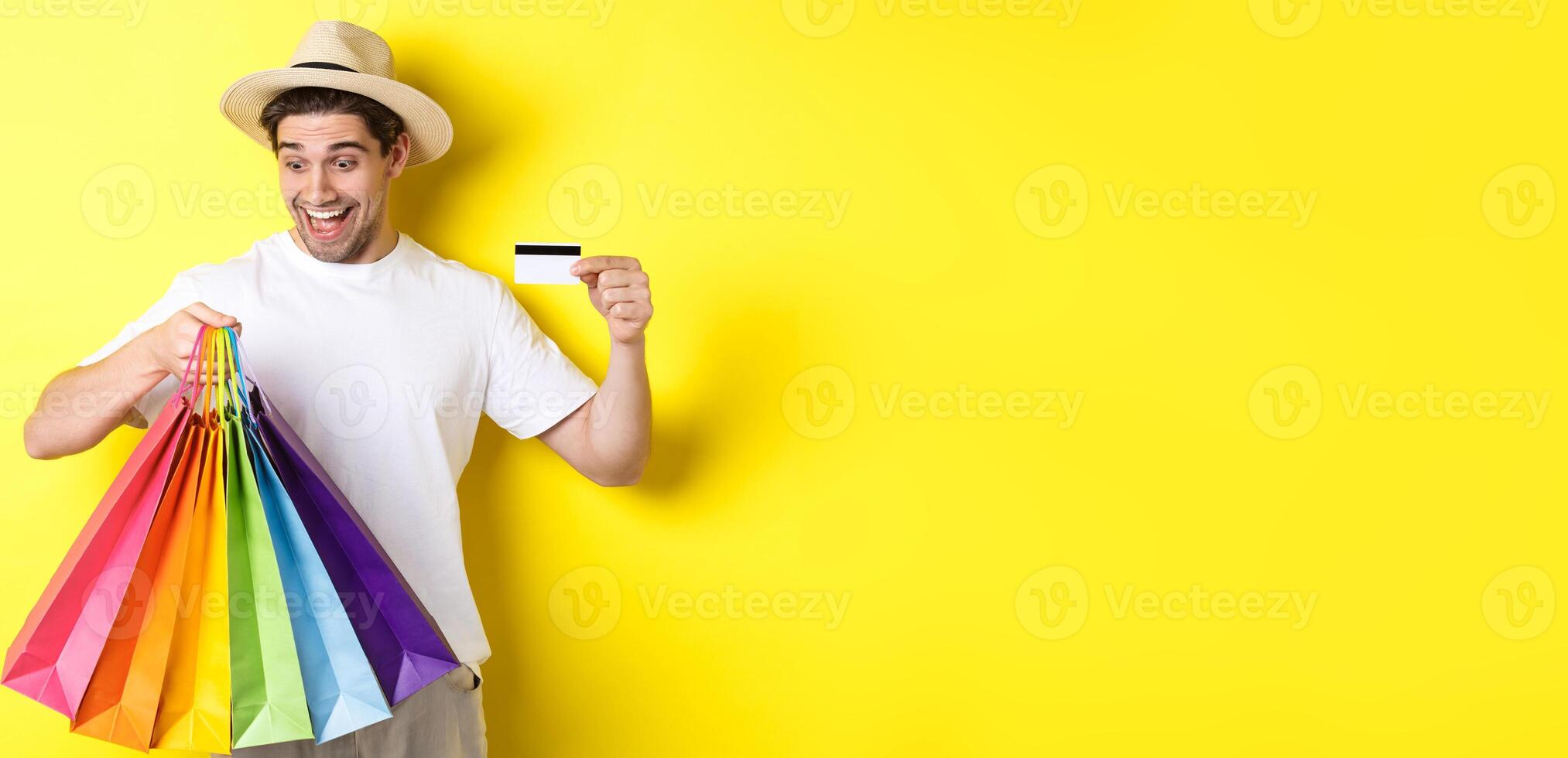 Concept of vacation and finance. Happy man shopper looking at shopping bags satisfied, showing credit card, standing against yellow background photo