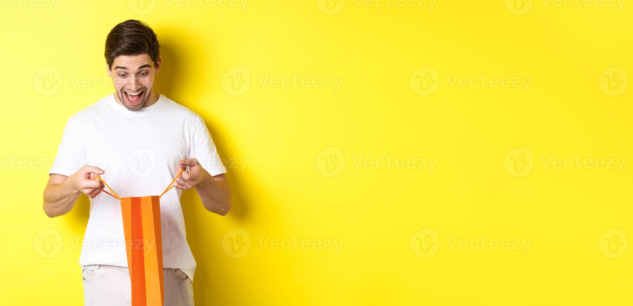 Excited guy open bag with gift, looking inside with amazement and happy face, standing against yellow background photo