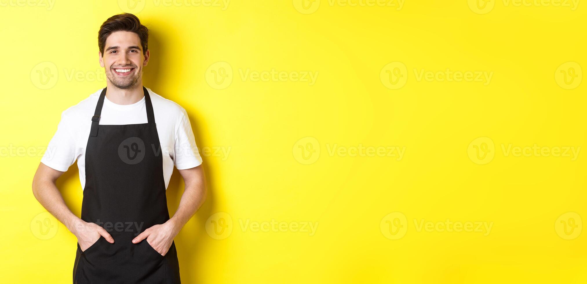 Happy barista in black apron looking at camera. Coffee shop owner wearing cafe uniform and smiling, standing over yellow background photo