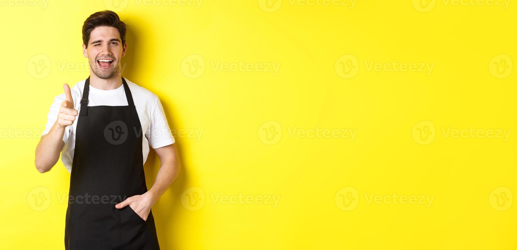 Handsome barista in black apron welcome guests to coffee shop, pointing finger gun and winking, standing against yellow background photo