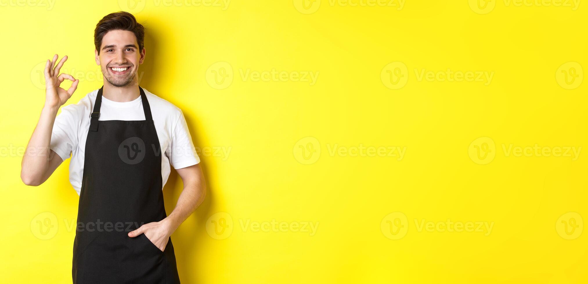 Young smiling barista in black apron showing okay sign, recommending coffee shop or restaurant, standing over yellow background photo