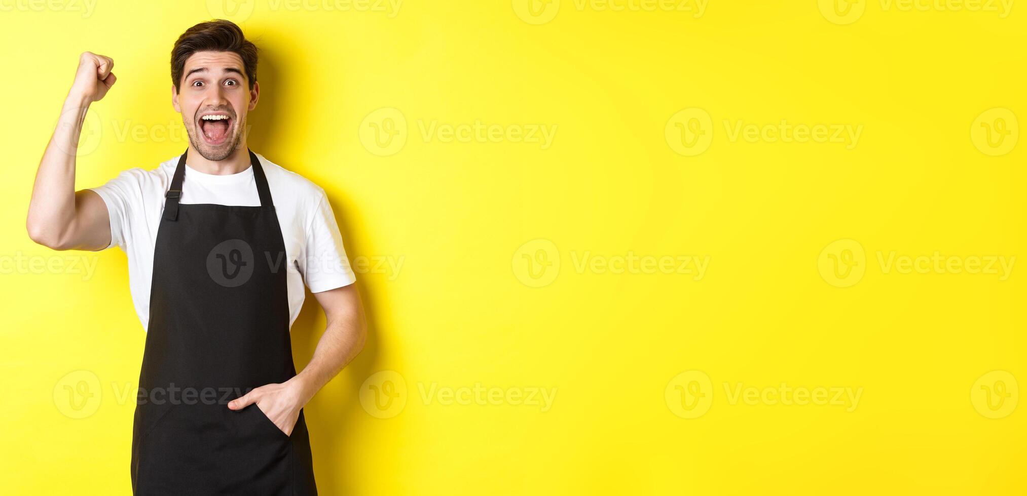 Cheerful seller making fist pump, rejoicing and triumphing, standing in black apron against yellow background photo