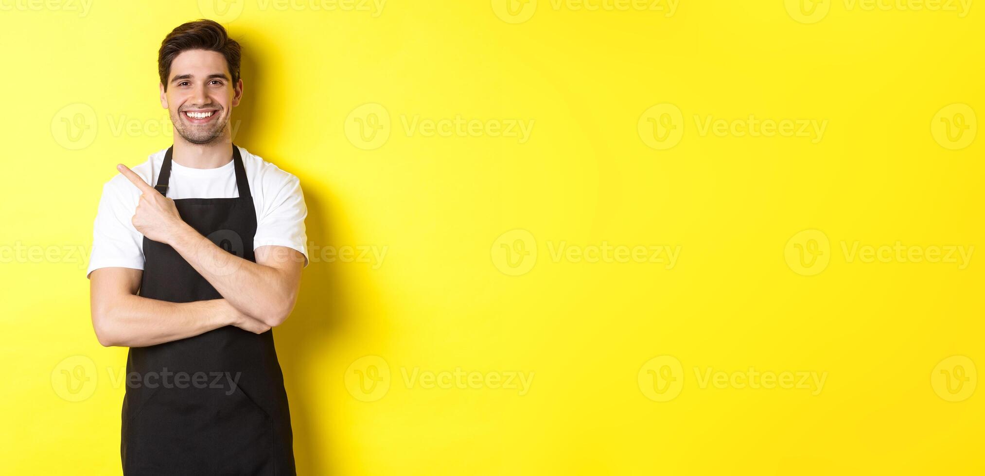 Happy barista pointing finger left and smiling, wearing black apron uniform, standing against yellow background photo