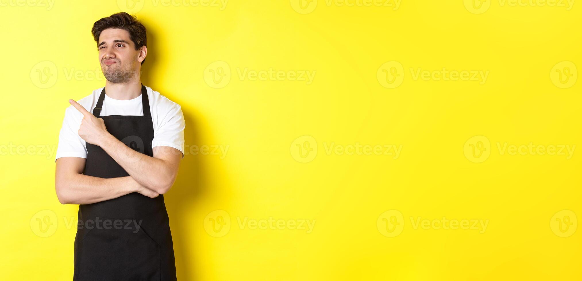 Skeptical male seller in black apron looking displeased, grimacing and pointing left at advertisement, standing over yellow background photo