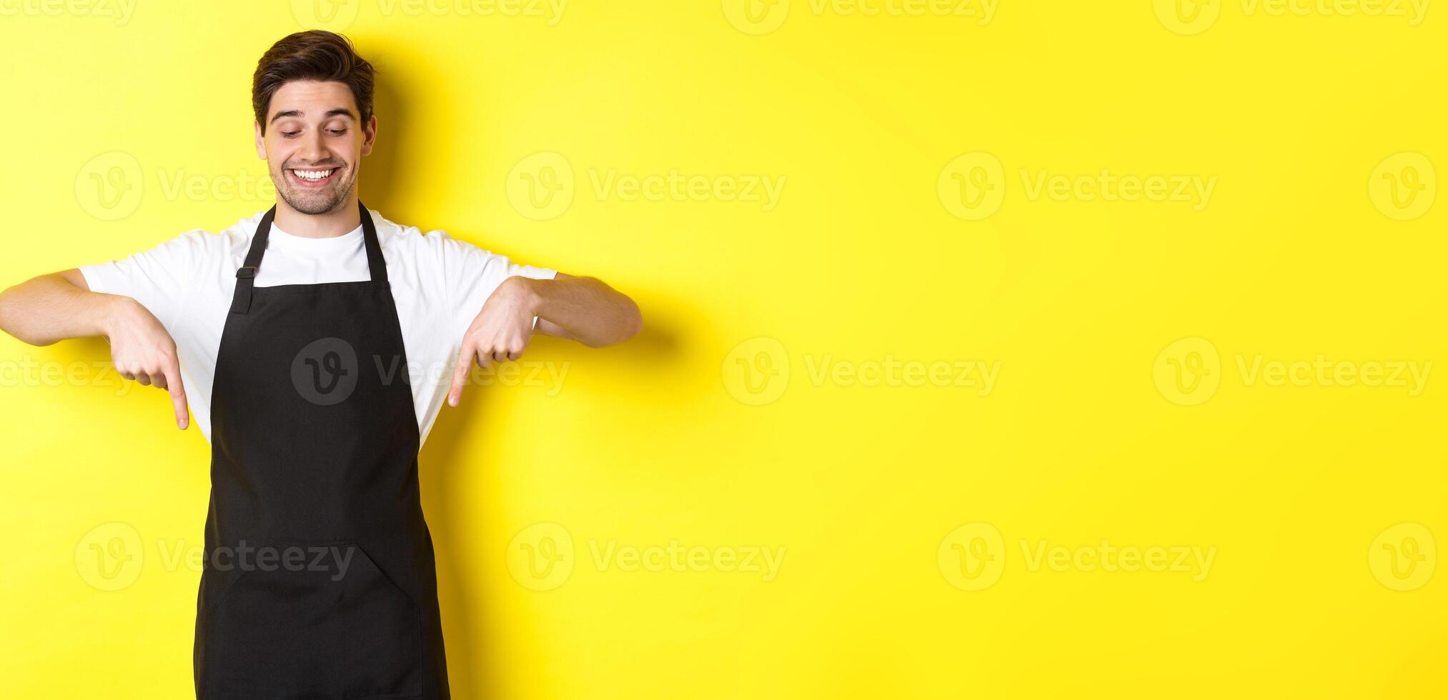 Happy coffee shop worker in black apron pointing fingers down, looking pleased and smiling, showing logo, standing over yellow background photo