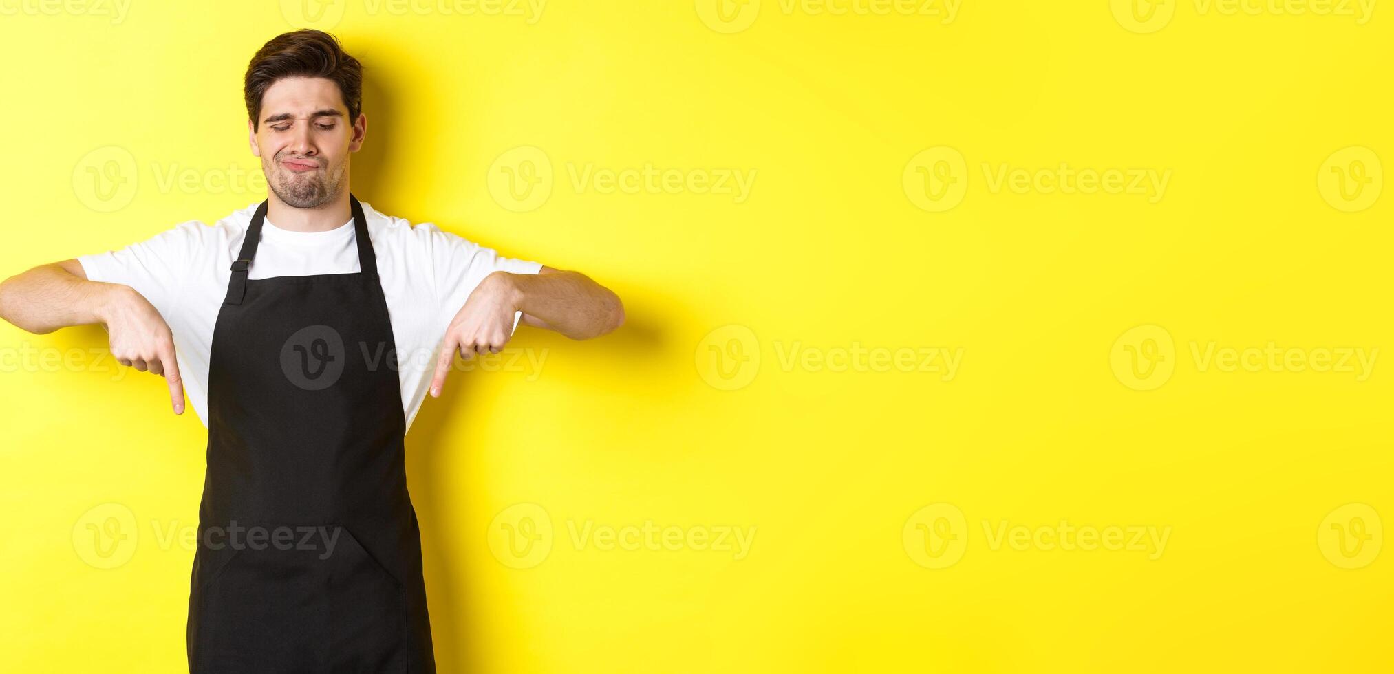 Doubtful barista complaining, pointing fingers down and grimacing displeased, standing in black apron against yellow background photo