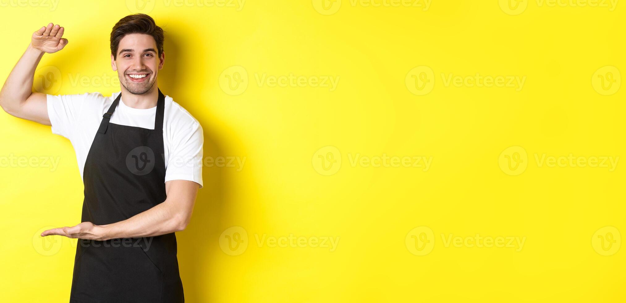 Smiling handsome barista showing something long or large, standing over yellow background photo