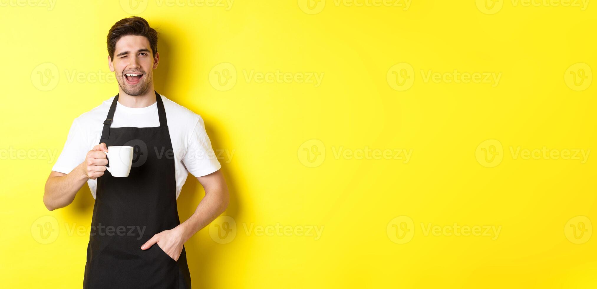 Cheerful barista in black apron serving coffee and winking, standing against yellow background photo