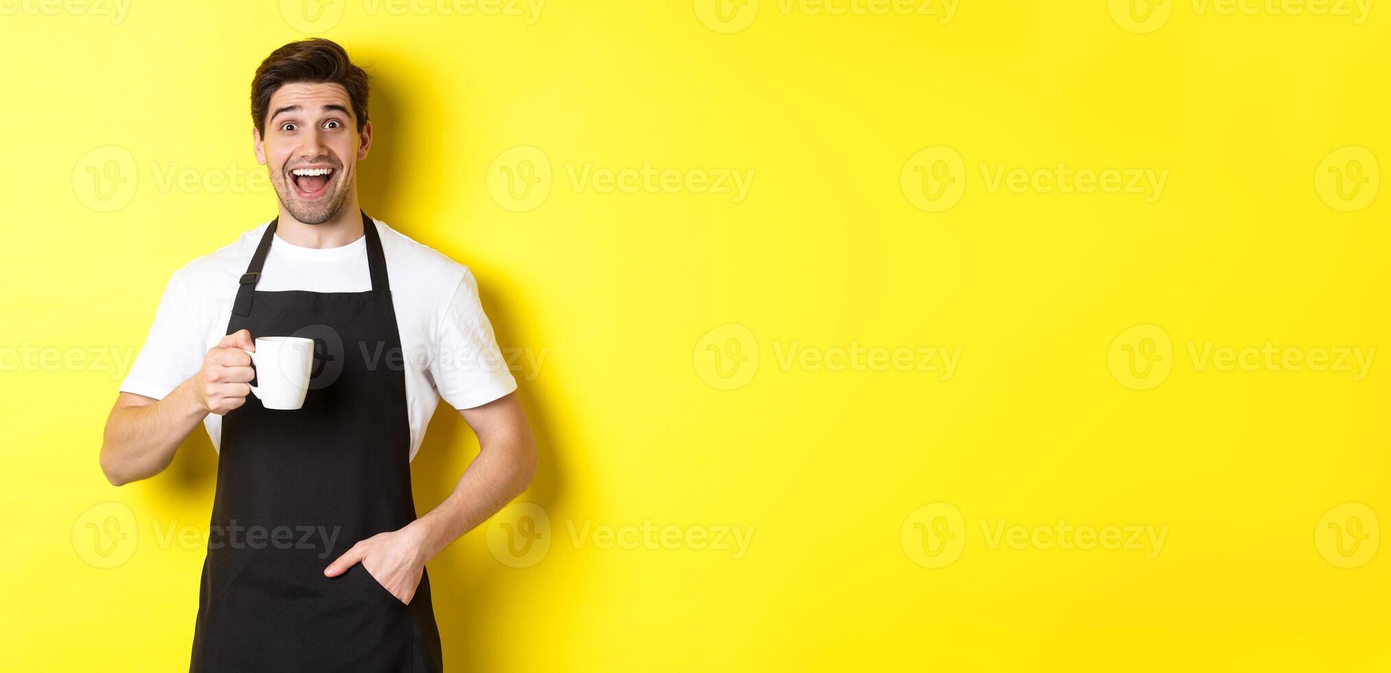 sonriente joven barista en negro delantal participación café taza, en pie terminado amarillo antecedentes foto