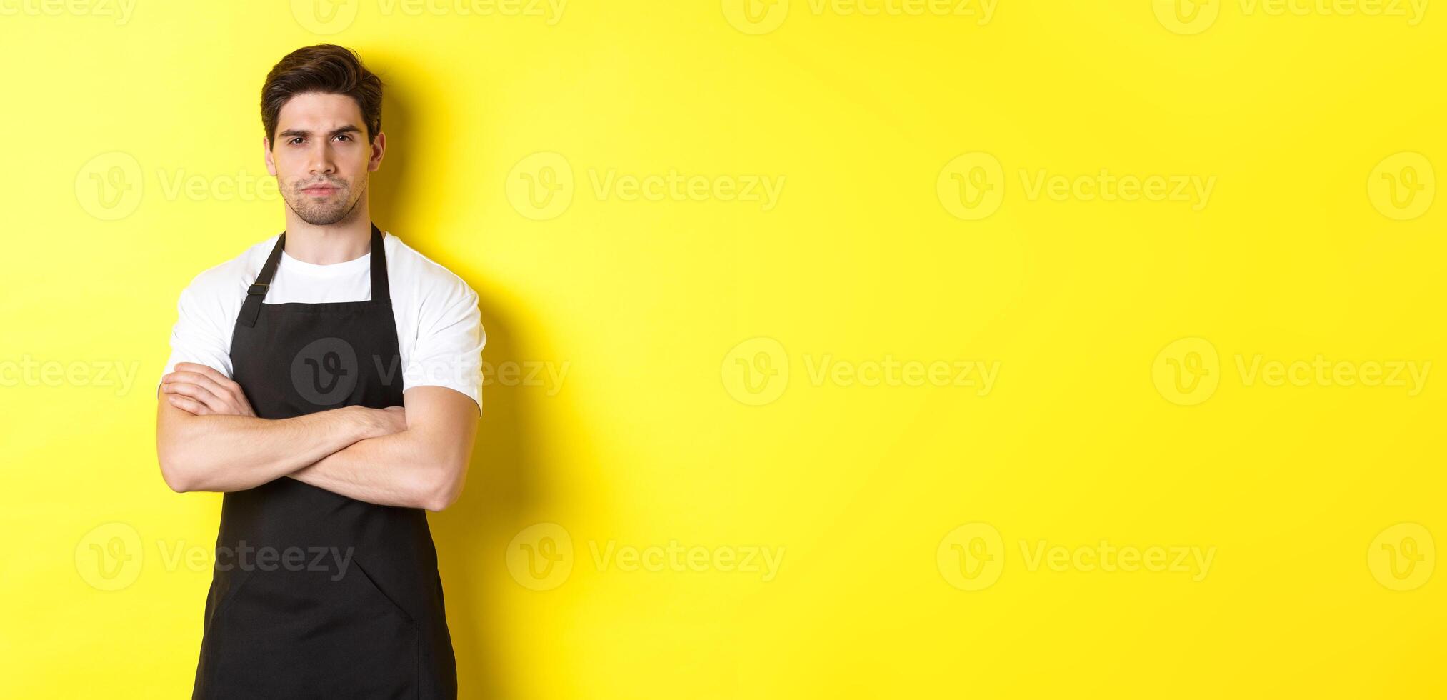 Suspicious barista squinting, cross arms on chest and looking at something with disbelief, standing over yellow background photo