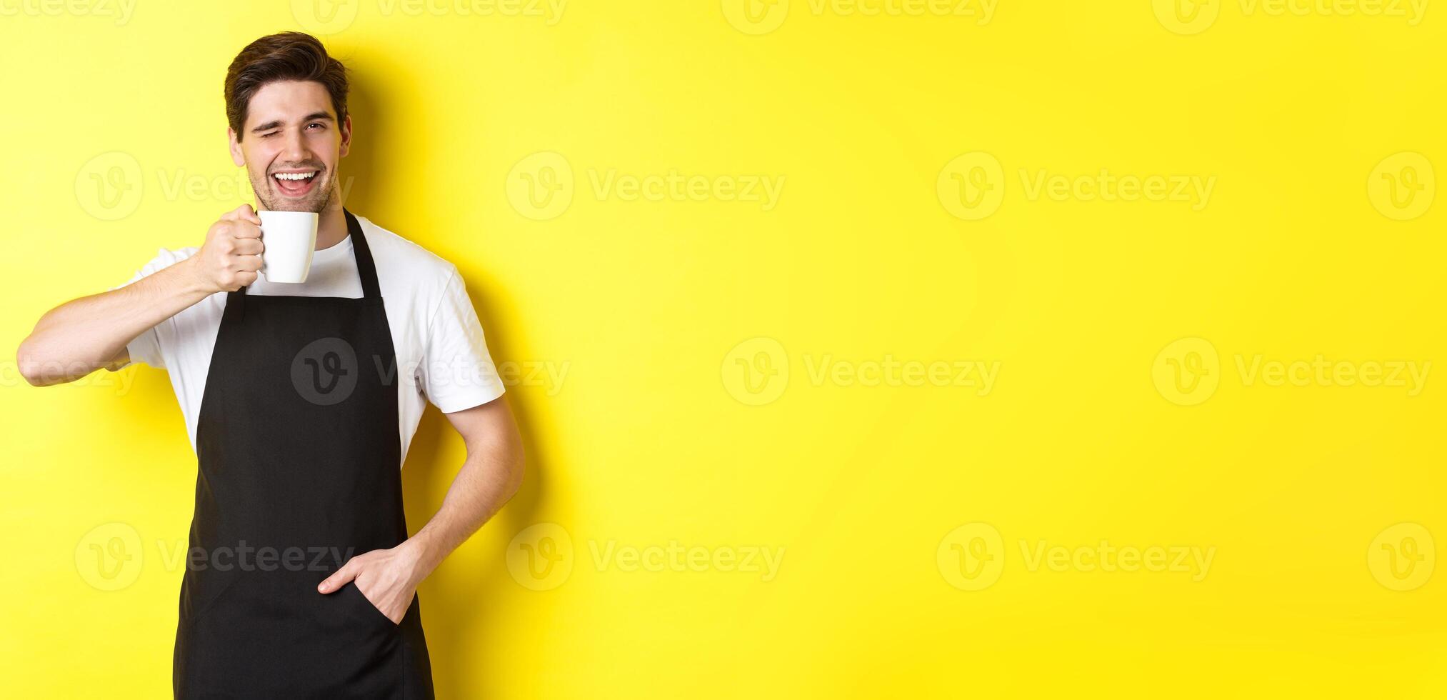 Handsome barista drinking cup of coffee and winking, inviting to cafe, standing over yellow background photo