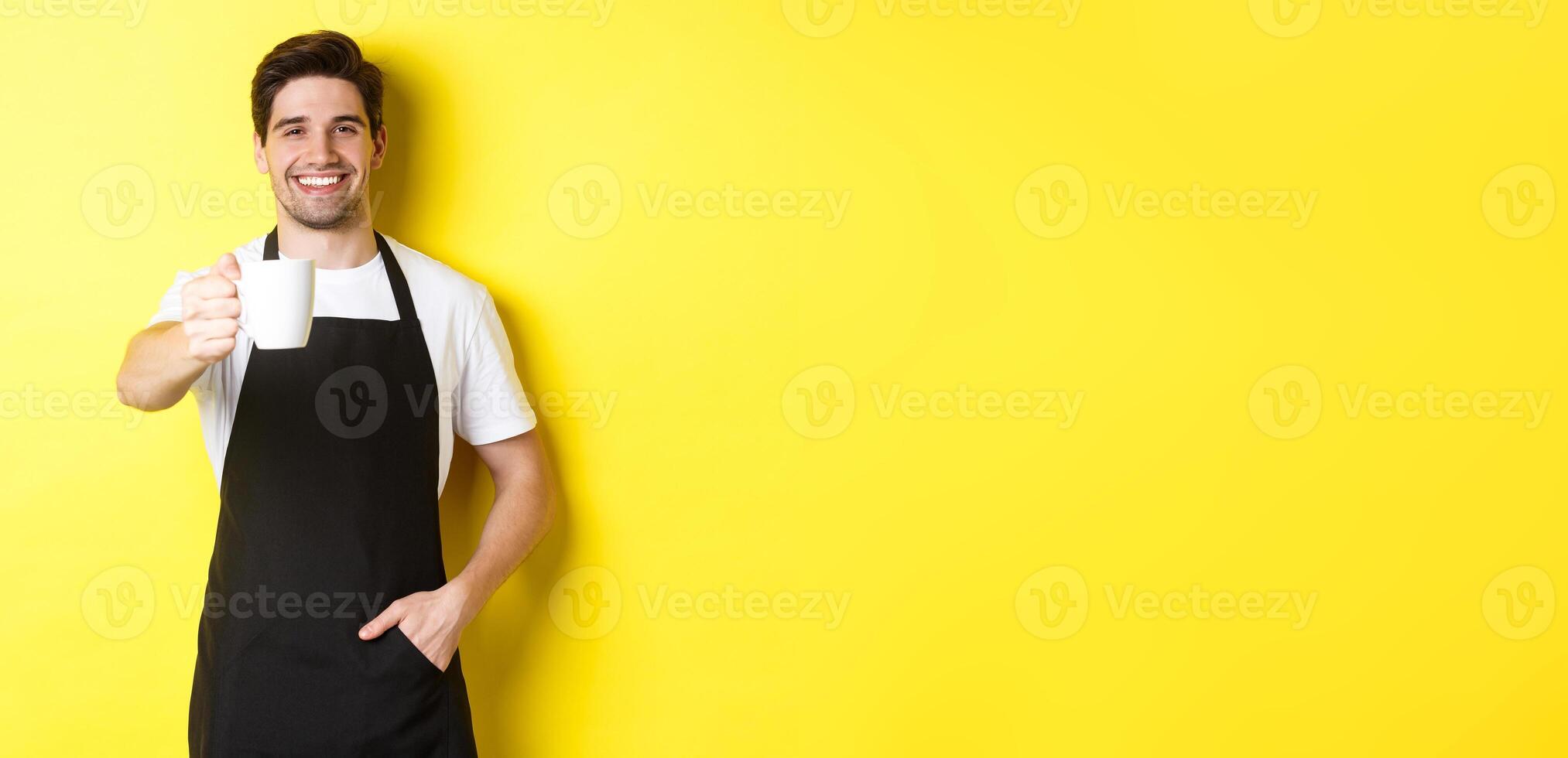 Handsome barista in black apron giving you cup of coffee and smiling, standing over yellow background photo