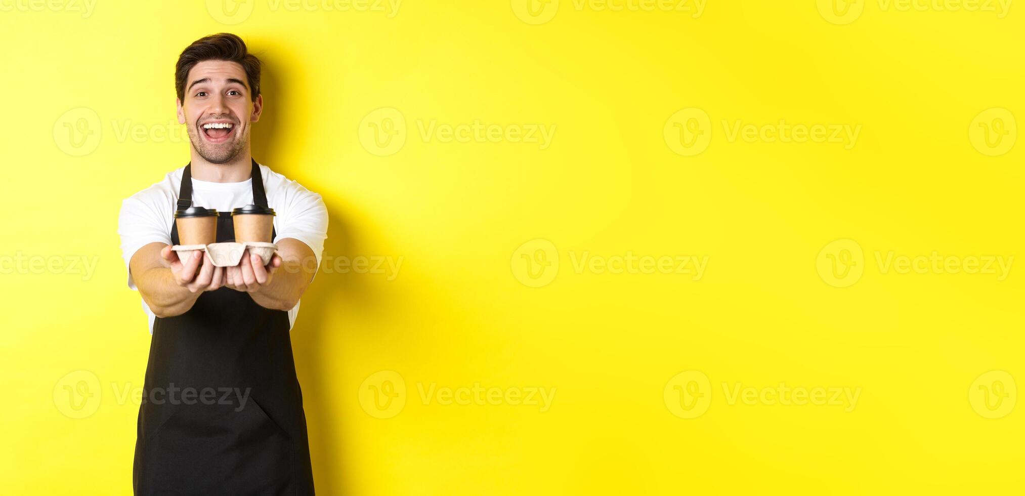 Friendly barista in black apron giving takeaway order, holding two cups of coffee and smiling, standing over yellow background photo
