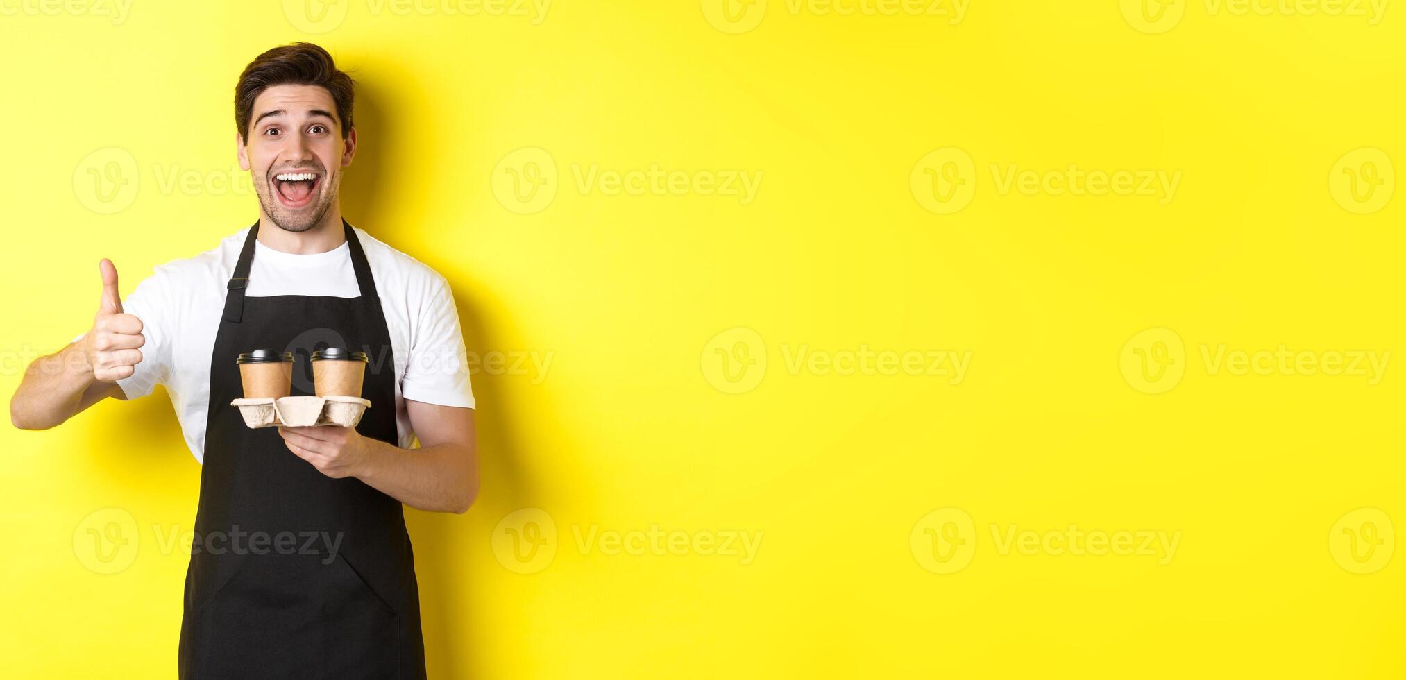 Cheerful barista recommend his cafe, holding takeaway coffee cups and showing thumbs up, standing against yellow background photo