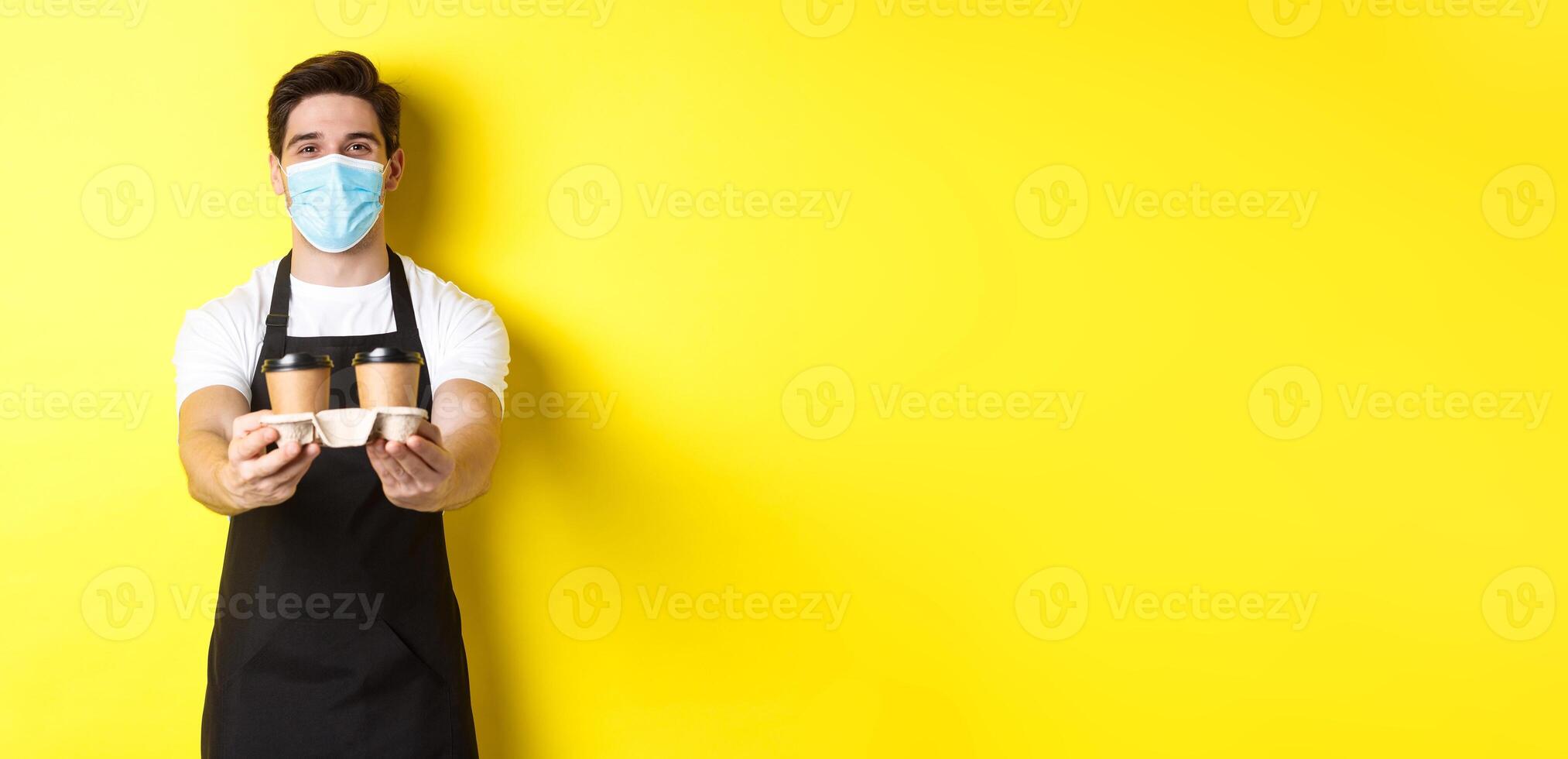 Concept of covid-19, cafe and social distancing. Barista in medical mask serving coffee in takeaway cups, standing in black apron against yellow background photo