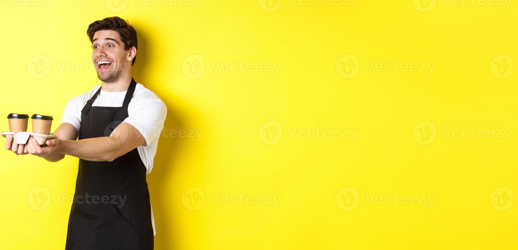Friendly barista in black apron giving takeaway order, holding two cups of coffee and smiling, standing over yellow background photo