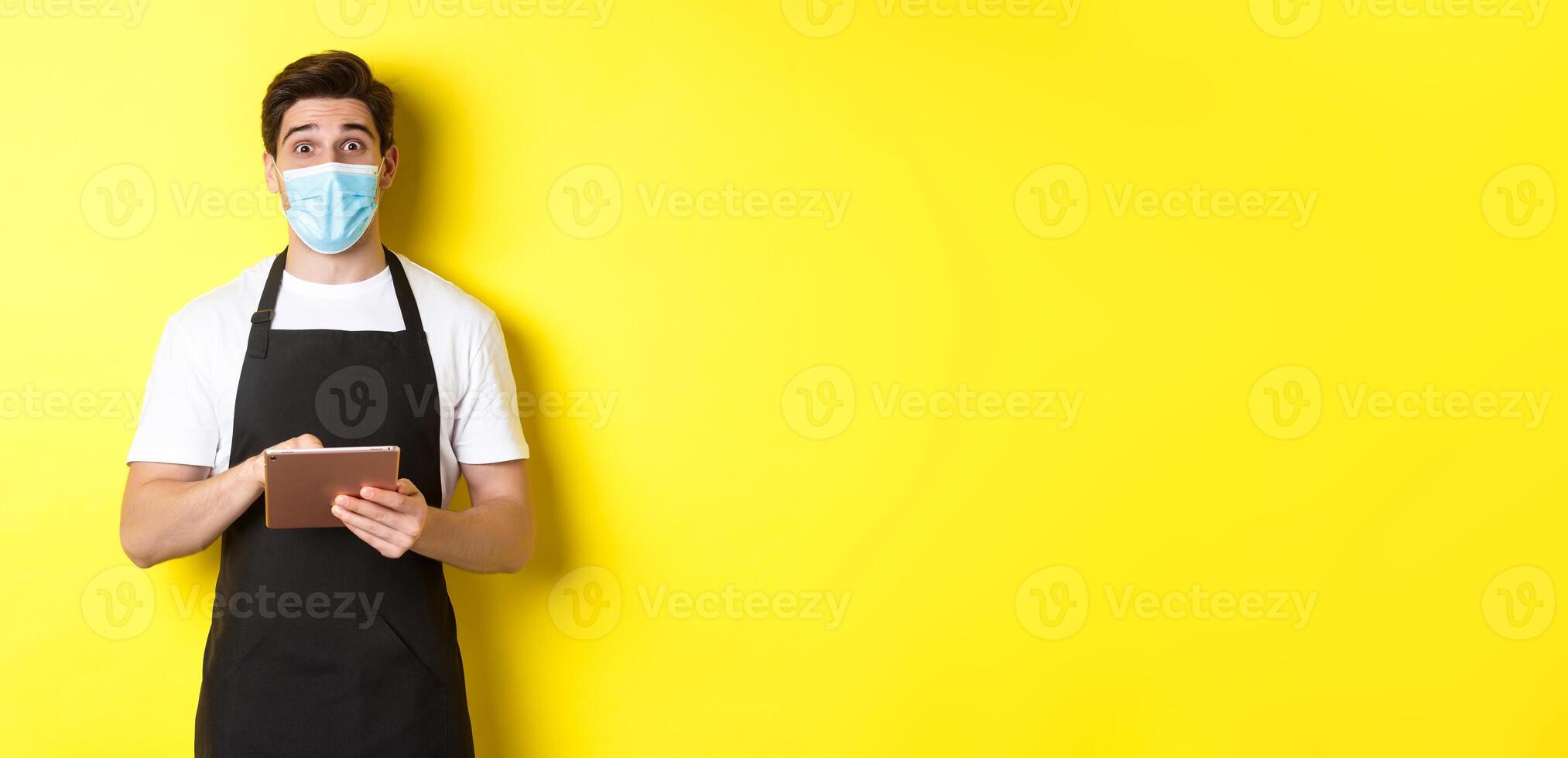 Concept of covid-19, small business and pandemic. Waiter in black apron and medical mask taking order, holding digital tablet, standing over yellow background photo