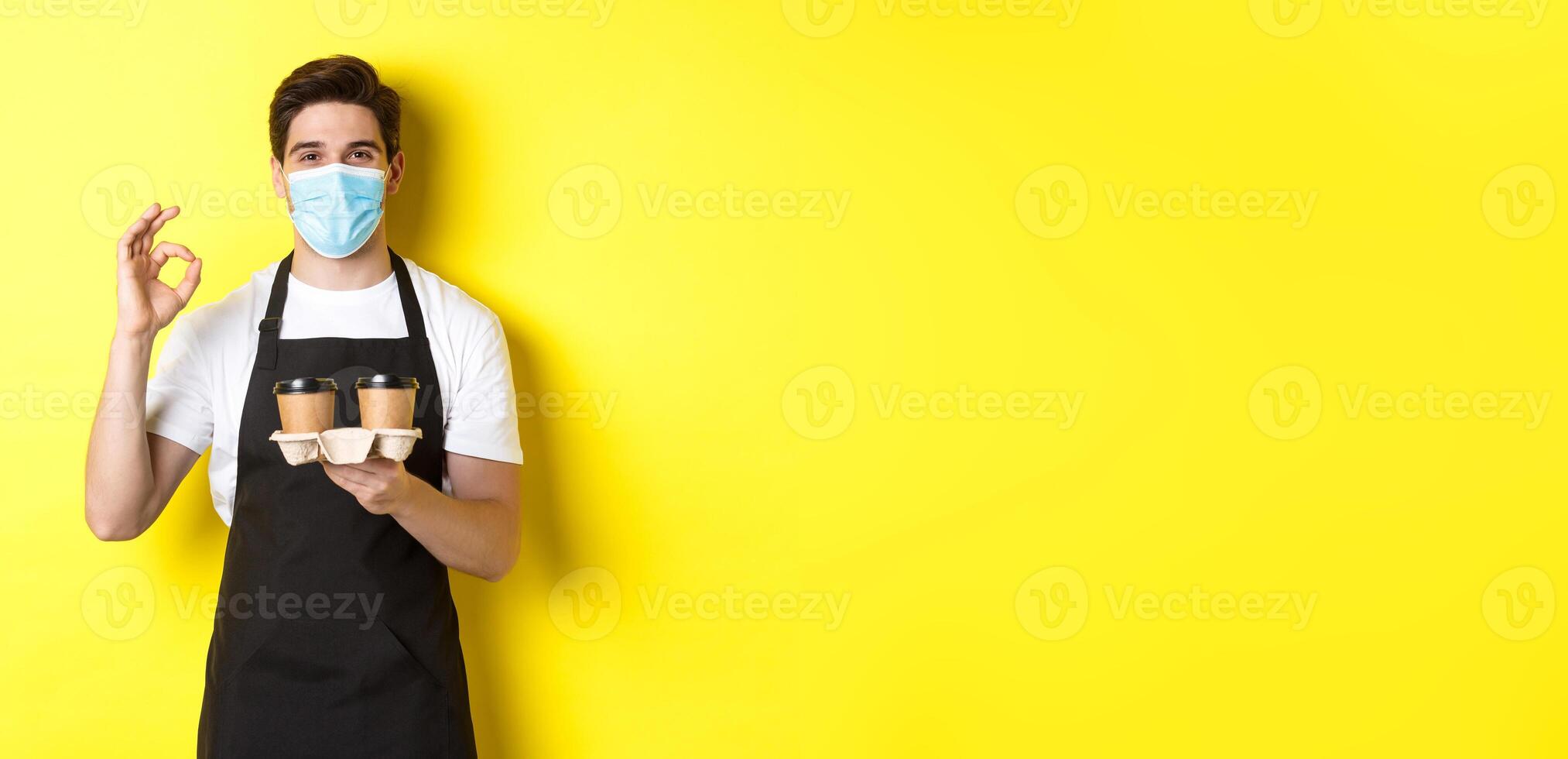 Concept of covid-19, cafe and social distancing. Barista in medical mask and black apron guarantee safety, holding takeaway cups of coffee and showing OK sign, yellow background photo