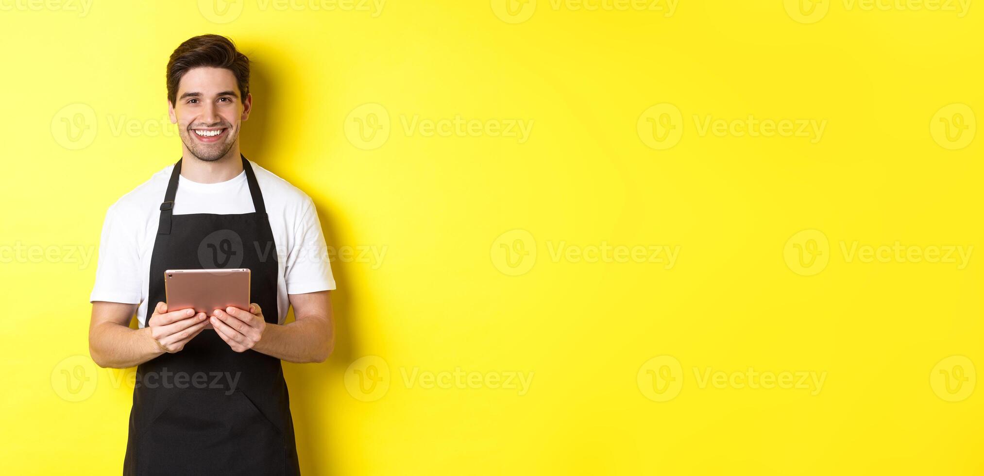 Waiter in black apron taking orders, holding digital tablet and smiling friendly, standing over yellow background photo