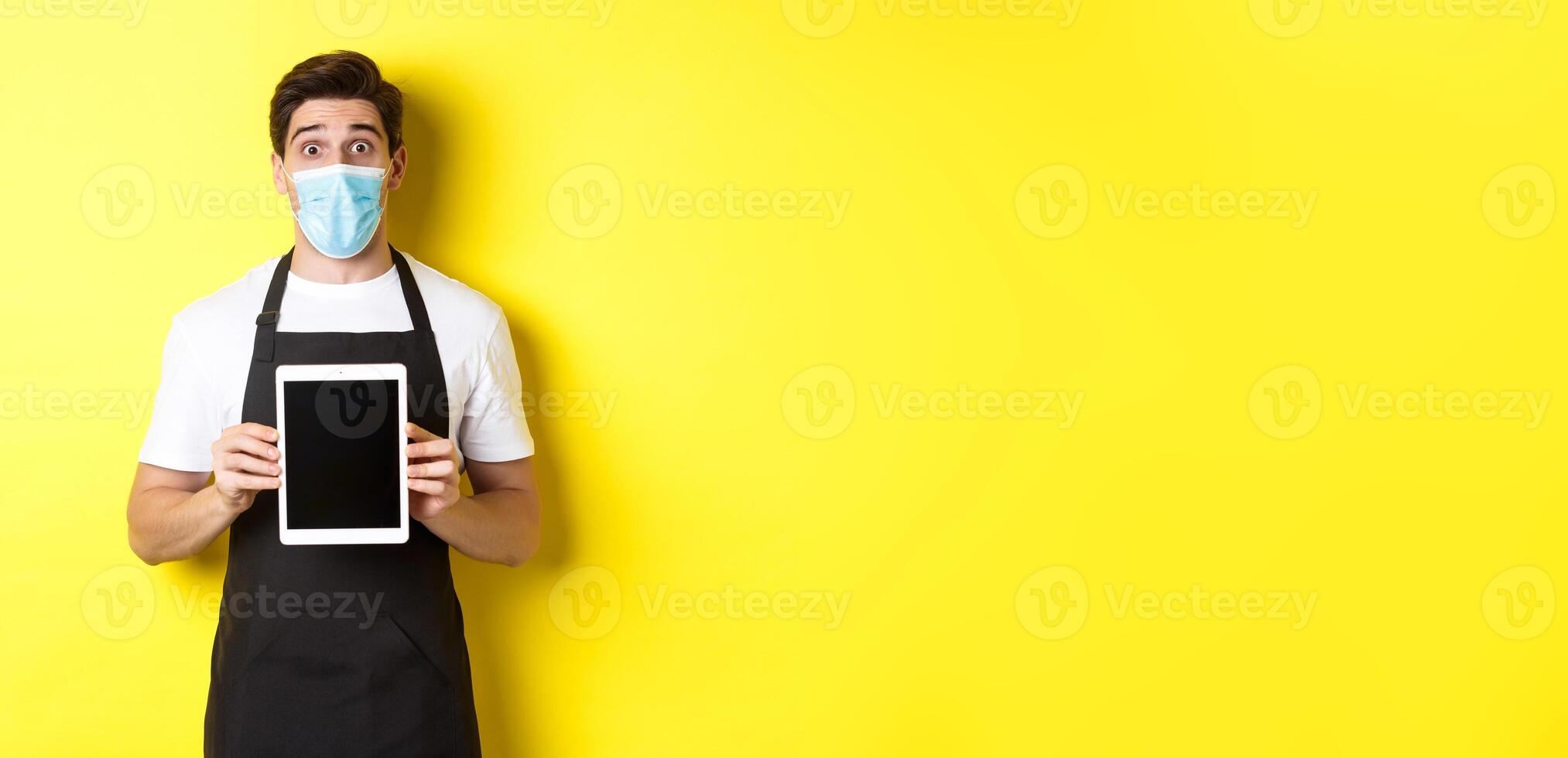 Concept of covid-19, small business and pandemic. Surprised waiter in black apron and medical mask showing tablet screen, standing over yellow background photo
