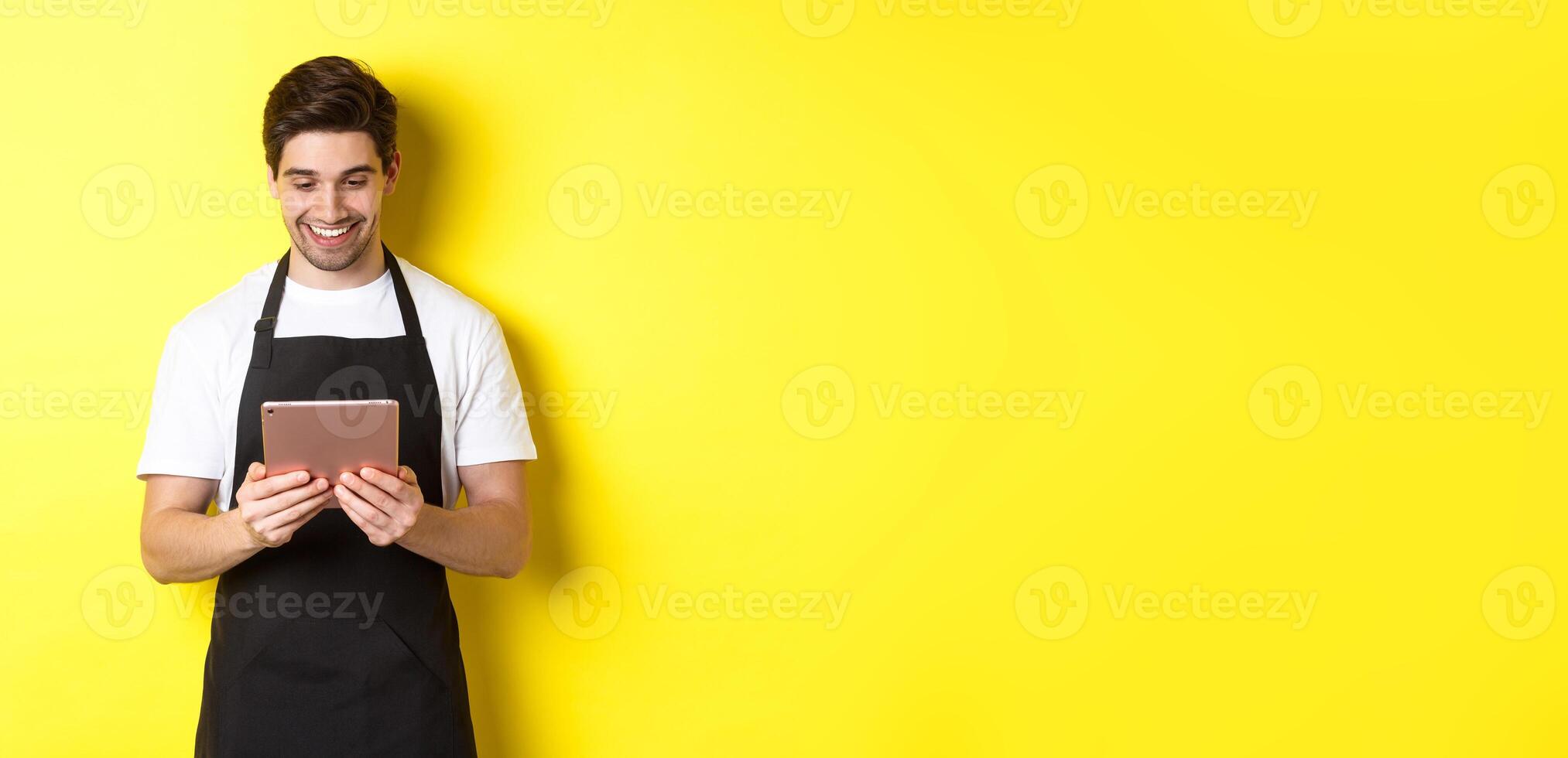 Seller in black apron looking at digital tablet screen, smiling pleased, standing over yellow background photo