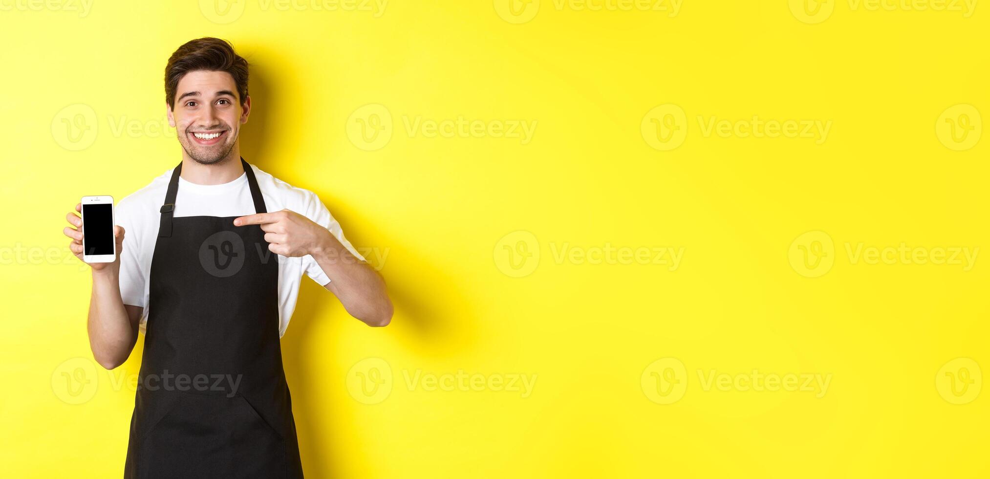 Handsome barista in black apron pointing finger at mobile screen, showing app and smiling, standing over yellow background photo