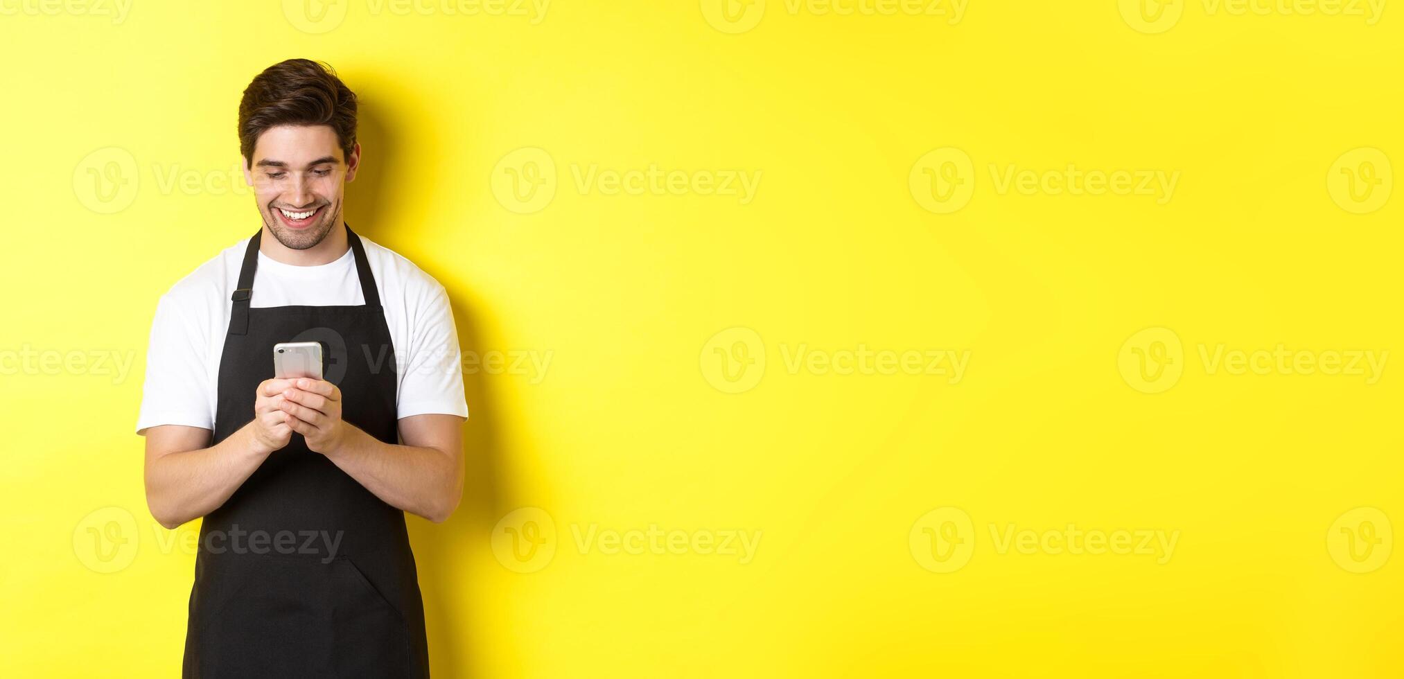 Waiter in black apron reading message on mobile phone, smiling happy, standing over yellow background photo