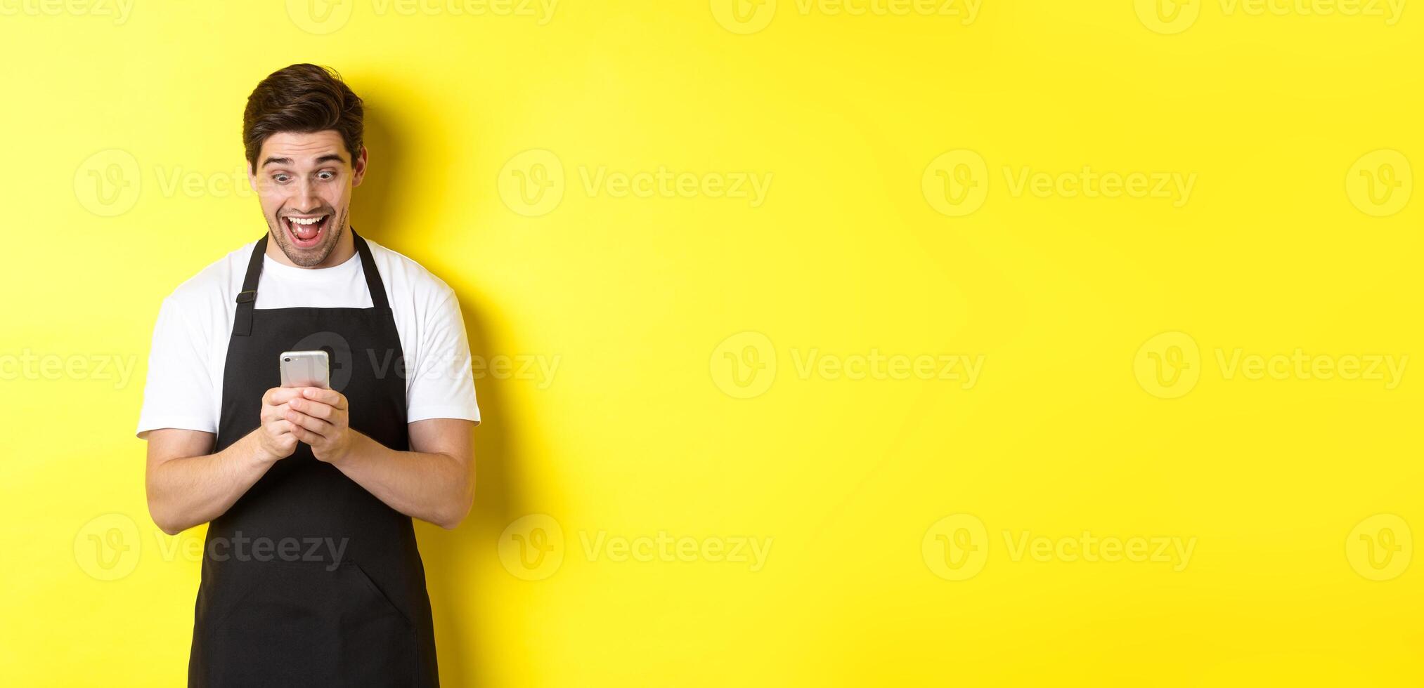 Barista looking surprised as reading message on mobile phone, standing in black apron against yellow background photo