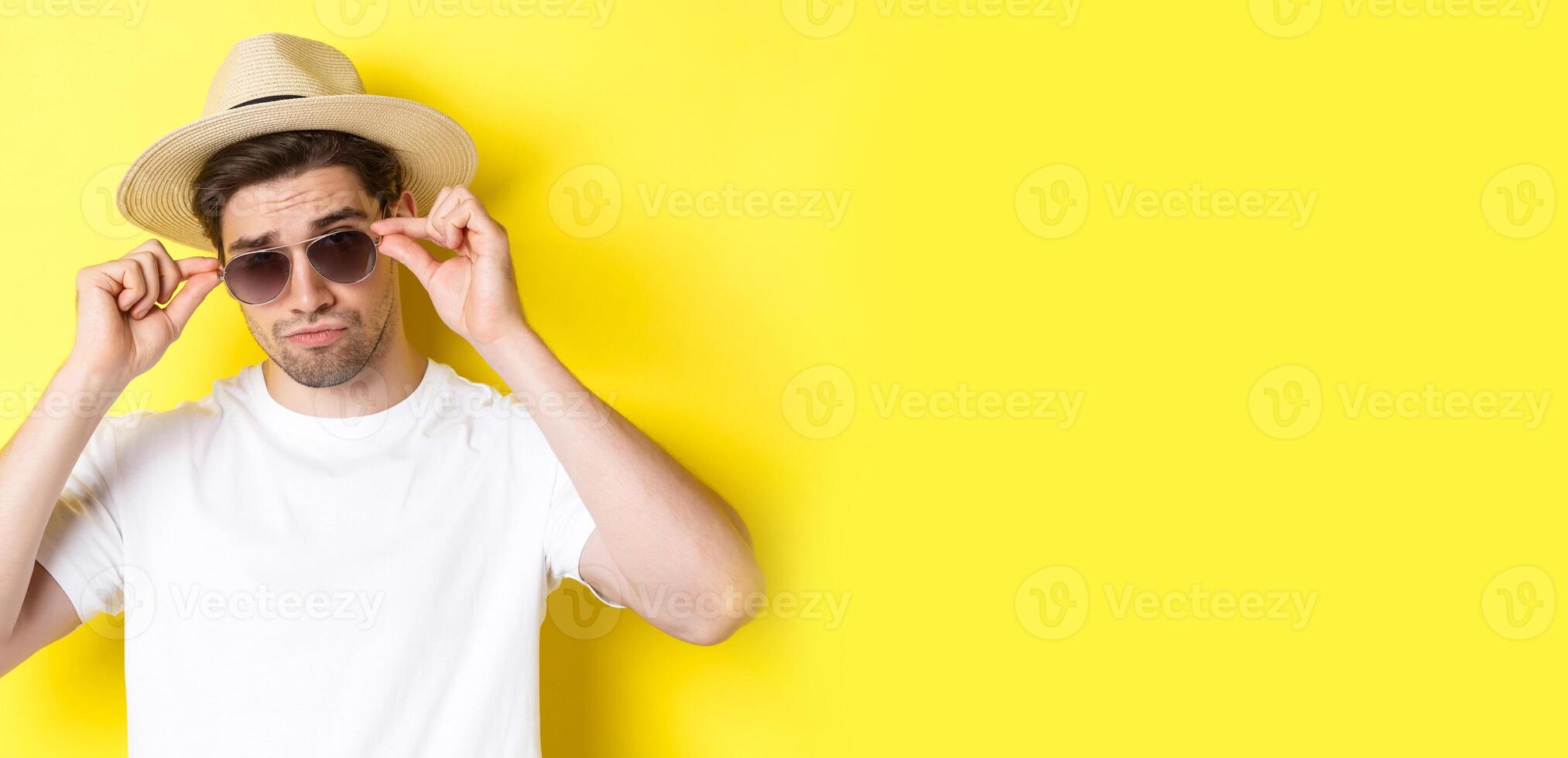 Concept of tourism and vacation. Close-up of cool tourist enjoying holidays on trip, wearing sunglasses with straw hat, yellow background photo