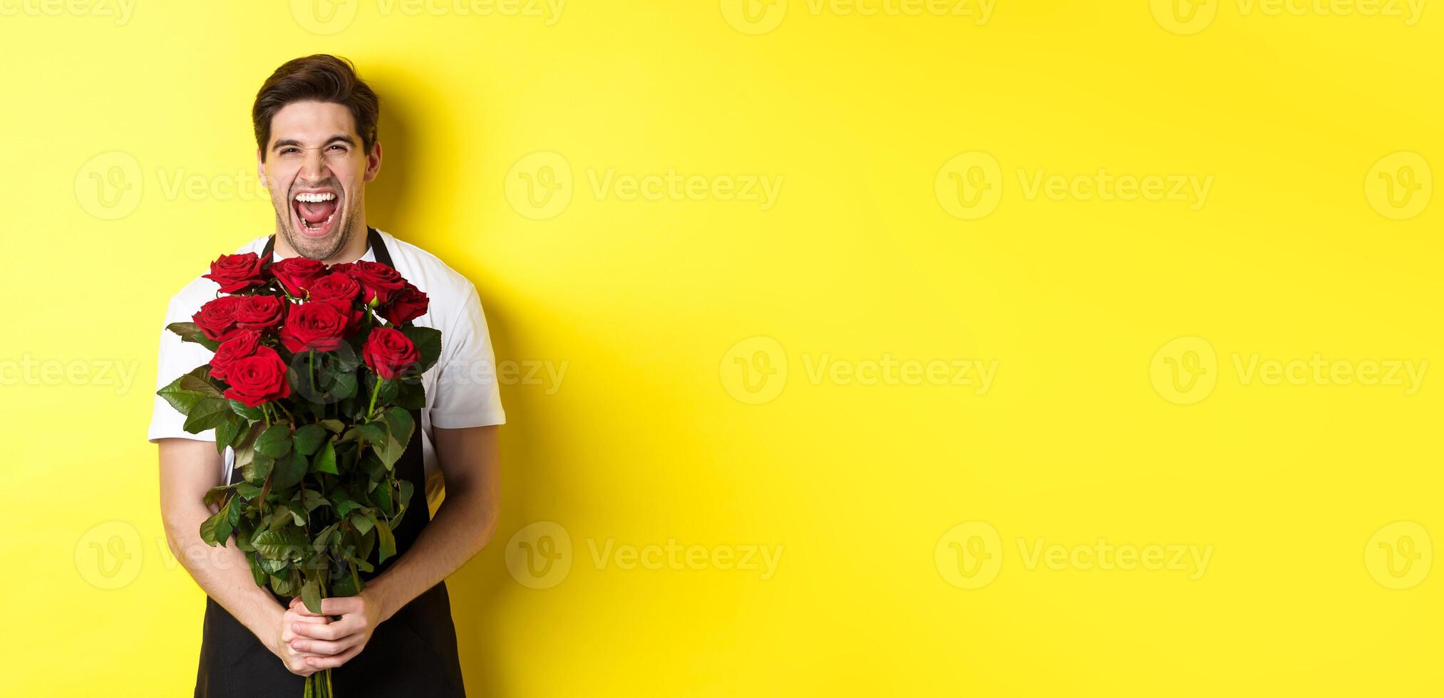 Funny young salesman in black apron holding bouquet of roses, florist laughing and standing over yellow background photo
