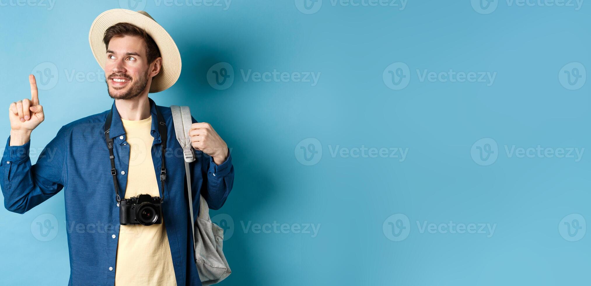 Handsome positive guy on vacation, wearing summer hat, looking and pointing up at logo, smiling pleased, standing with camera and backpack on blue background photo
