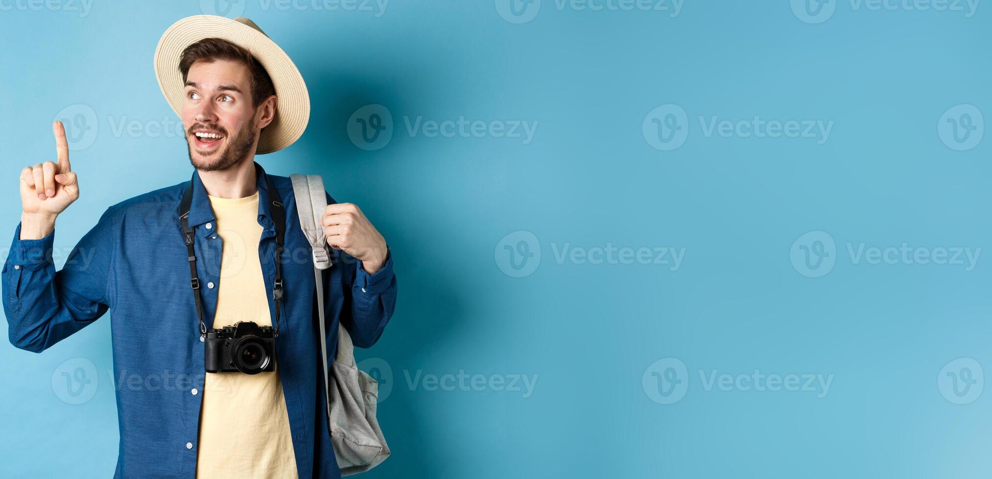 Handsome tourist looking and pointing up, travelling on summer vacation with backpack and camera, standing on blue background photo
