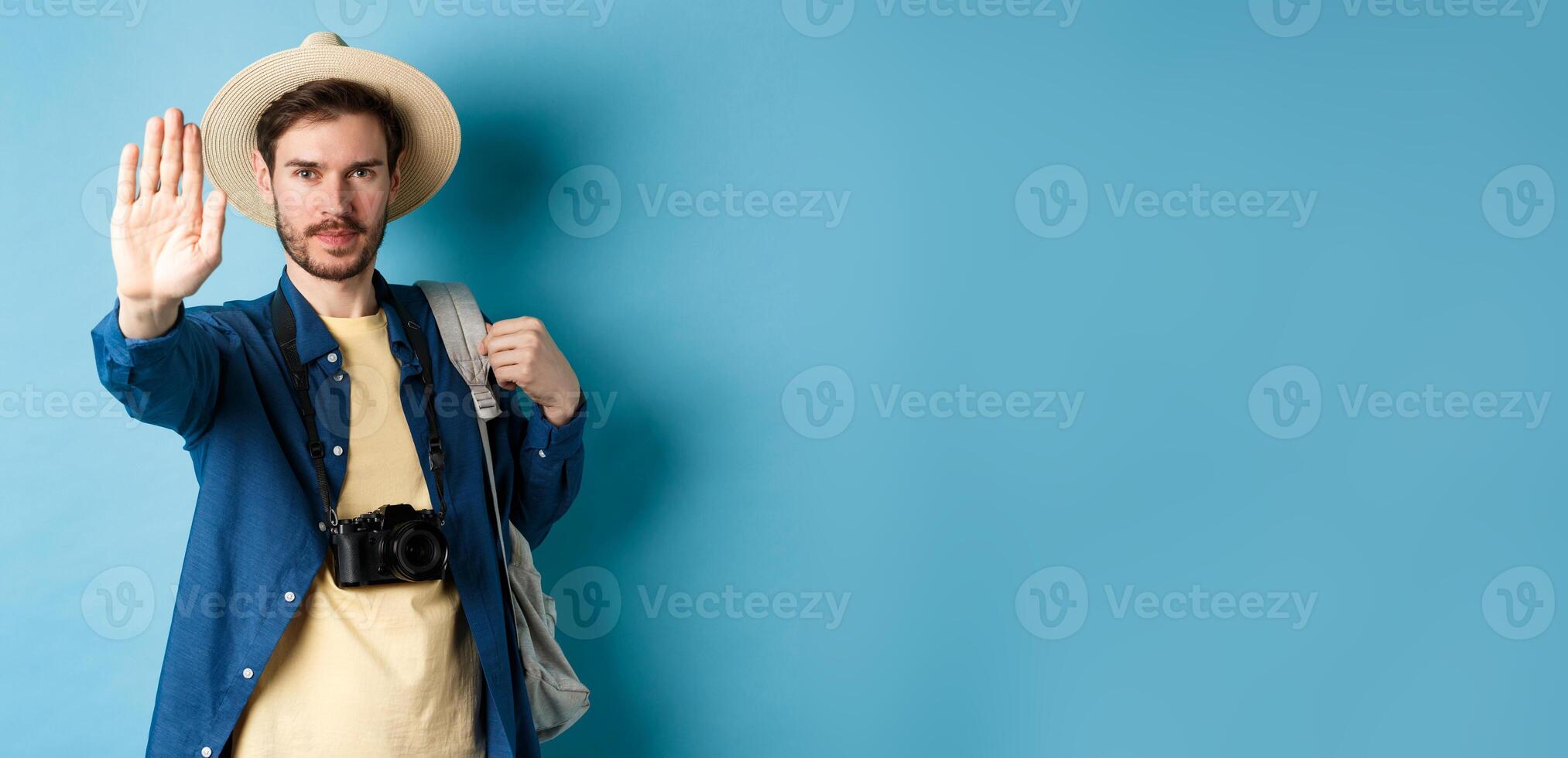 Serious male tourist warn you, stretch out hand to show stop gesture. Man on vacation forbid something, refusing or prohibiting action, standing with backpack on blue background photo