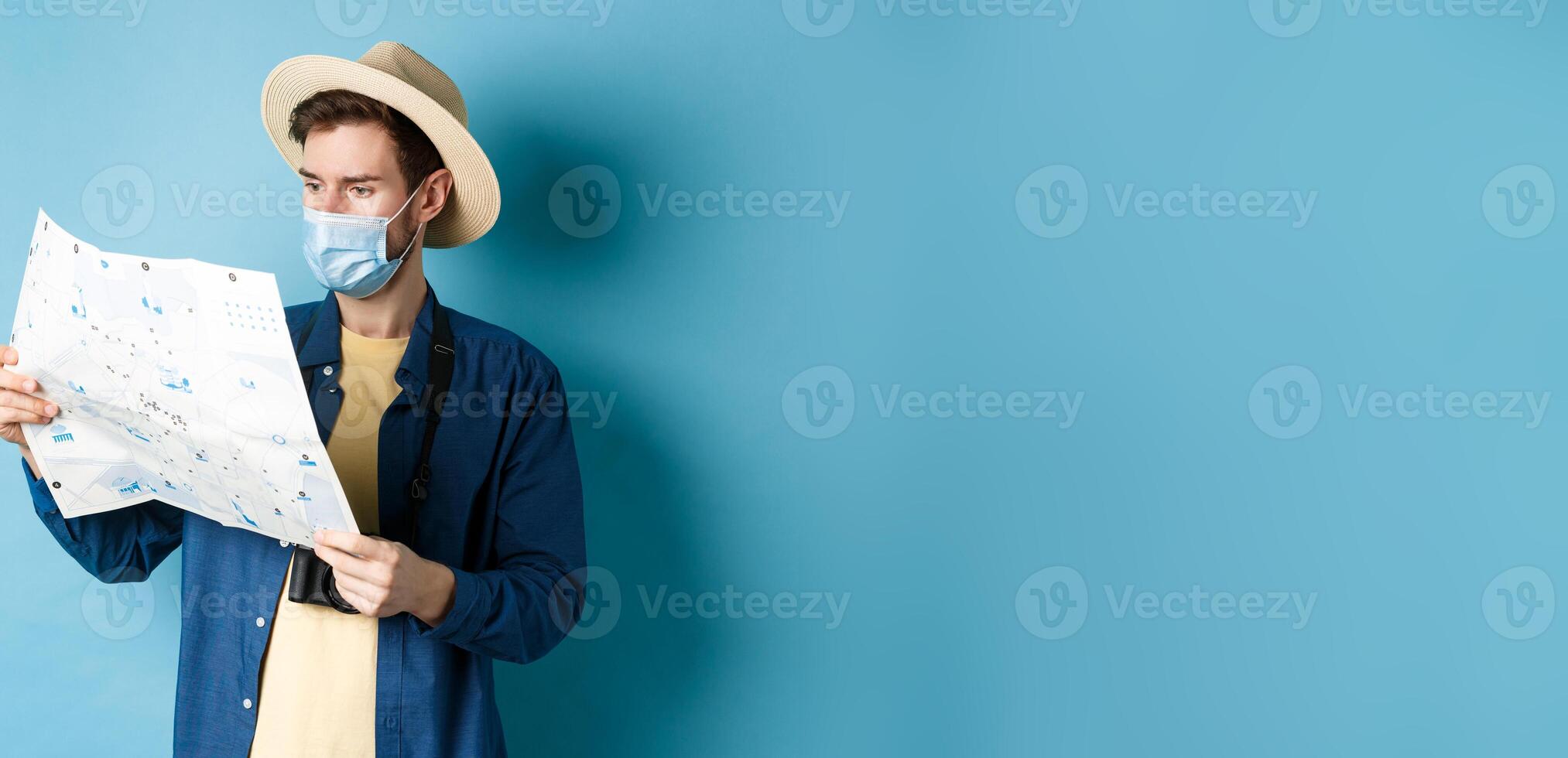 Covid-19, pandemic and travel concept. Handsome young guy in face mask and summer hat looking at tourist map, travelling on vacation, standing on blue background photo