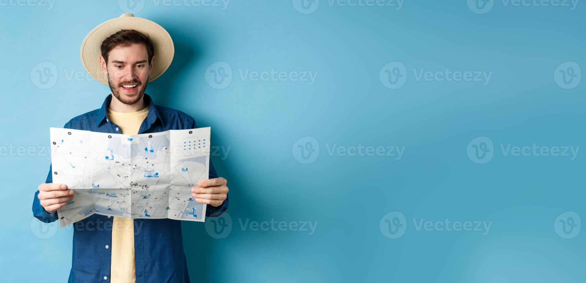 Cheerful tourist in summer hat planning travel route on vacation, looking at sighseeing map and smiling excited, standing on blue background photo