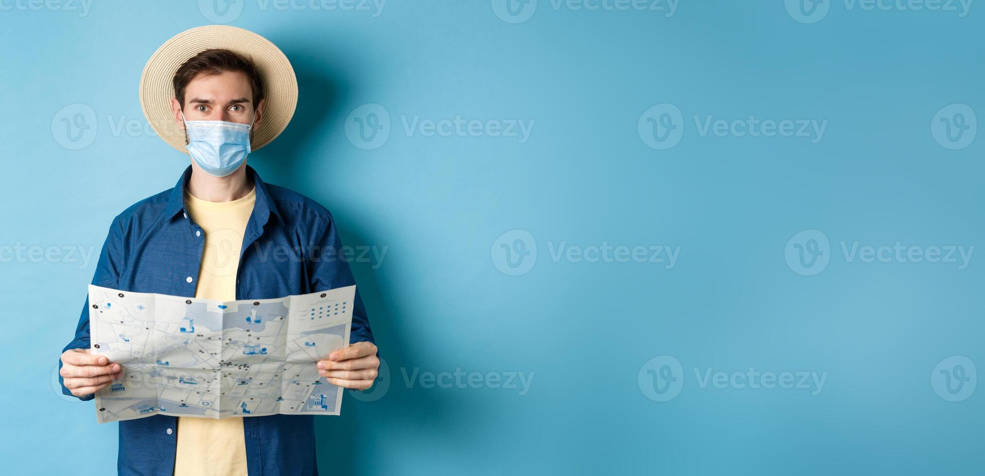 Covid-19, pandemic and travel concept. Happy tourist on summer vacation wearing medical mask and straw hat, holding road map, standing on blue background photo