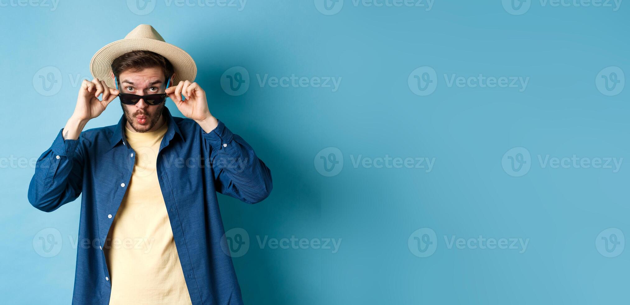 Portrait of tourist takes-off sunglasses and saying wow, staring impressed at camera, checking out cool thing, standing in summer hat on blue background photo