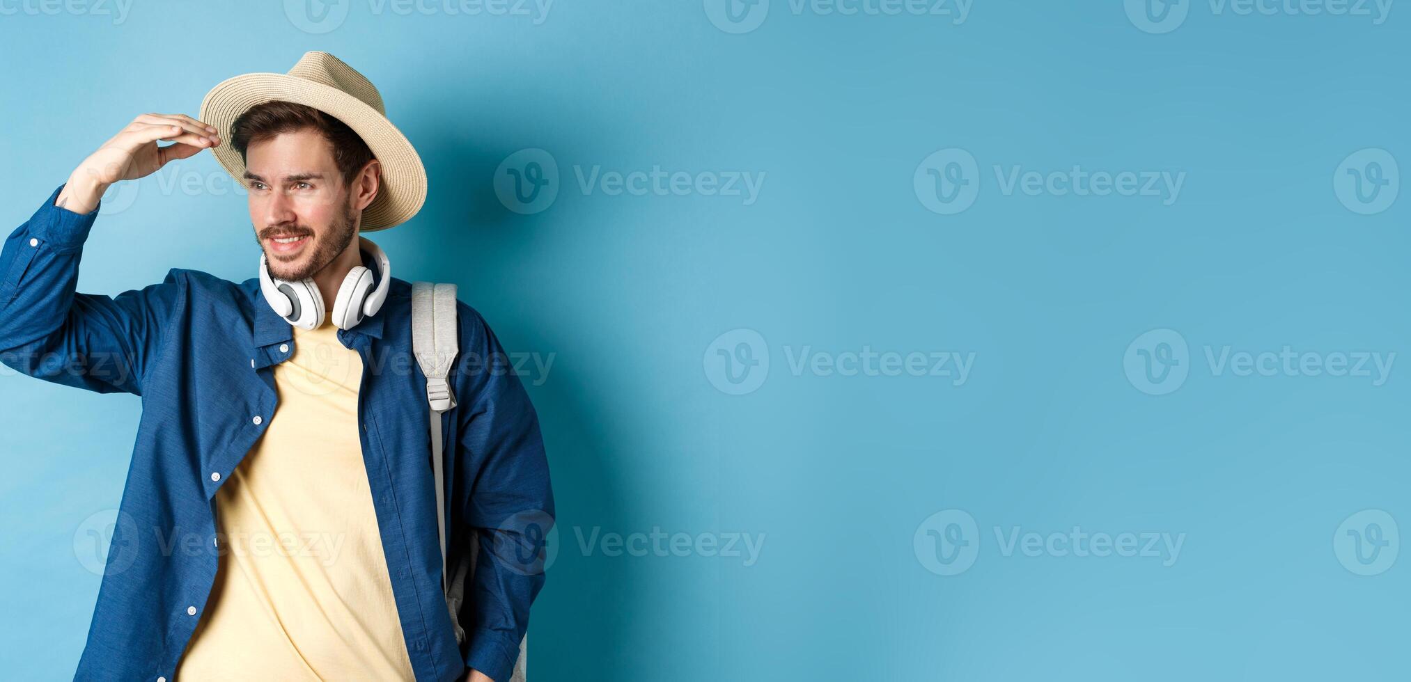 Handsome tourist in straw hat, covering face from sun and looking at distance aside, smiling pleased, travelling on summer vacation, standing over blue background photo