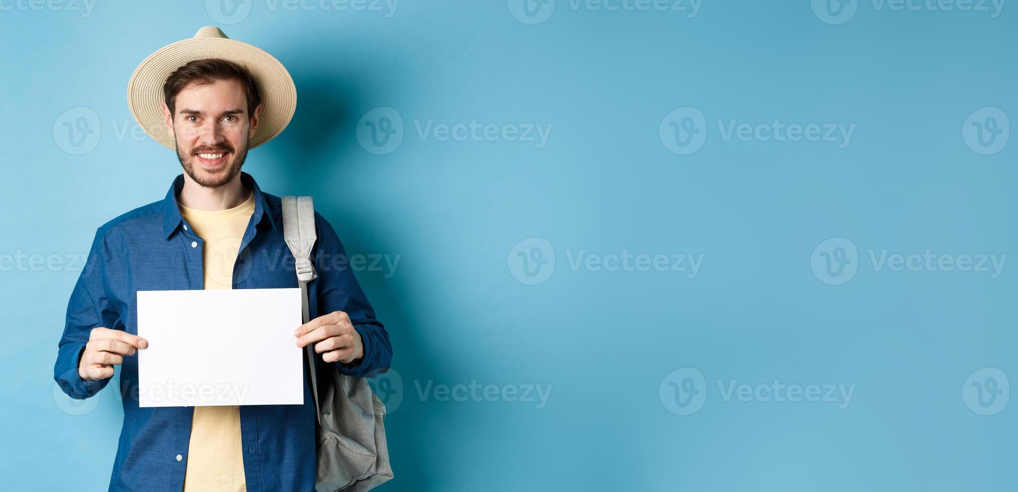 Happy tourist with backpack showing empty piece of paper, smiling at camera, standing on blue background. Concept of summer holiday and vacation photo