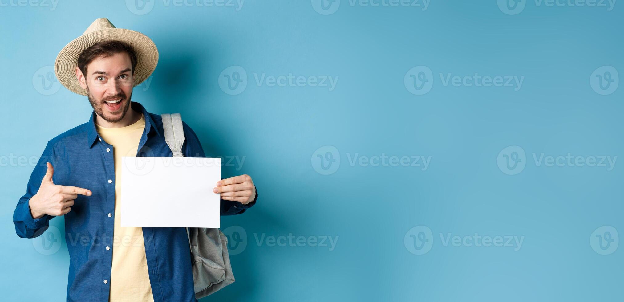 Handsome tourist smiling and pointing at empty piece of paper, wearing straw hat and backpack. Concept of summer vacation photo