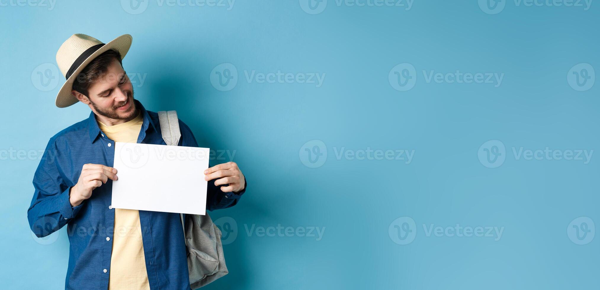 Happy tourist in straw hat smiling and looking at piece of paper for your logo, standing on blue background photo