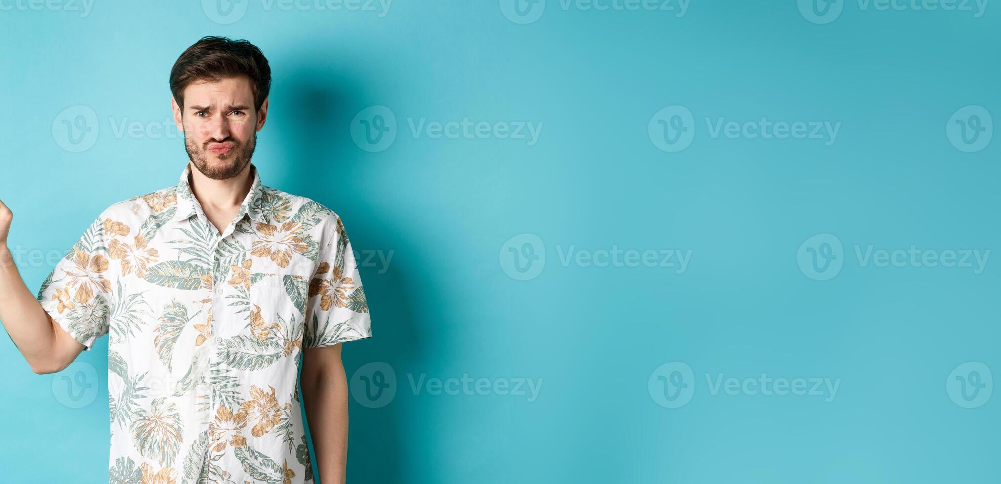 Disappointed tourist cringe and frown, pointing finger aside at empty space, complaining on bad thing, standing in hawaiian shirt on blue background photo